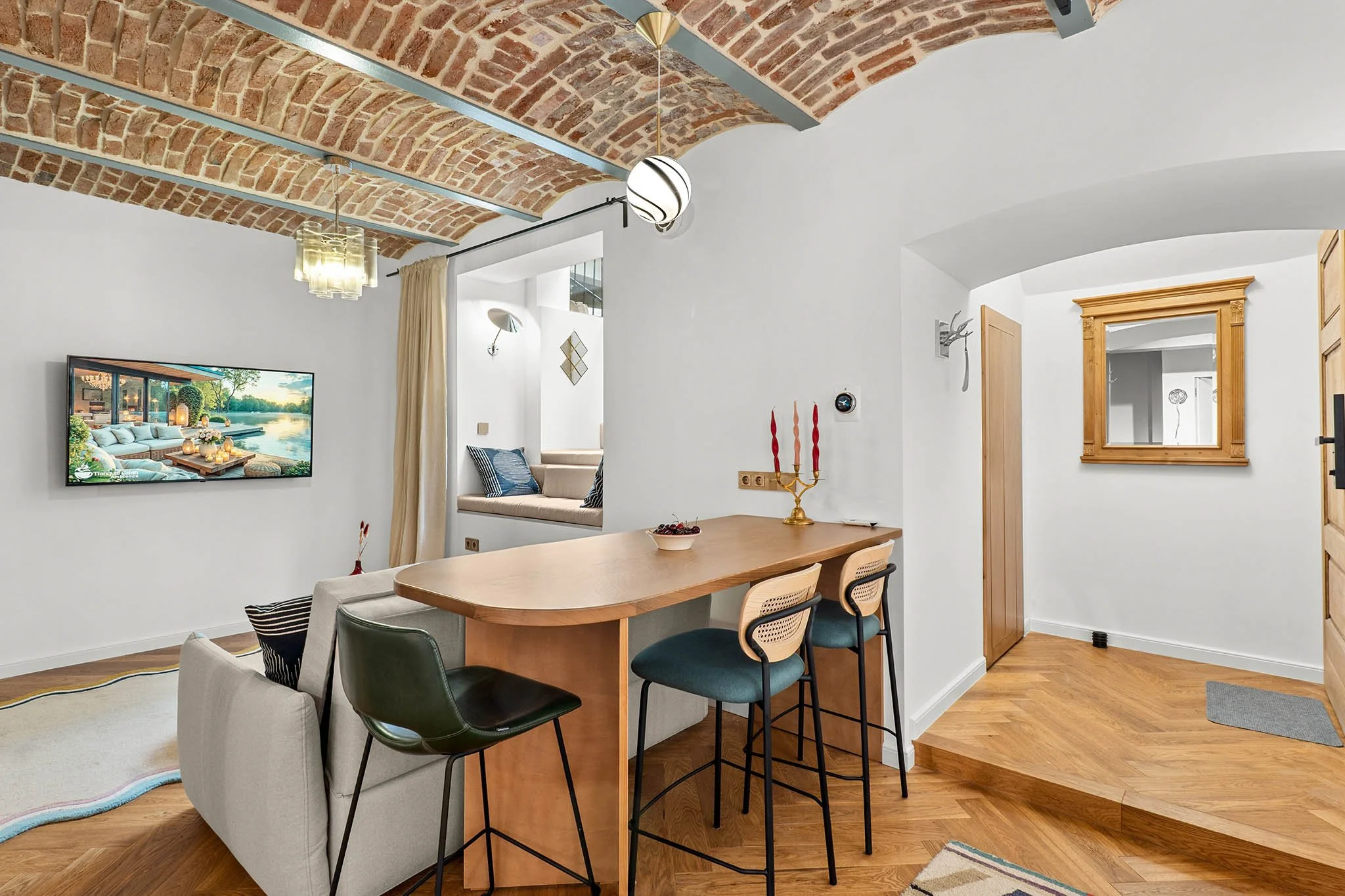 Living room with arched brick ceiling, white walls, wooden floor, TV mounted on wall, cozy window seat, beige couch, wooden dining table with colorful candles, and modern chairs.