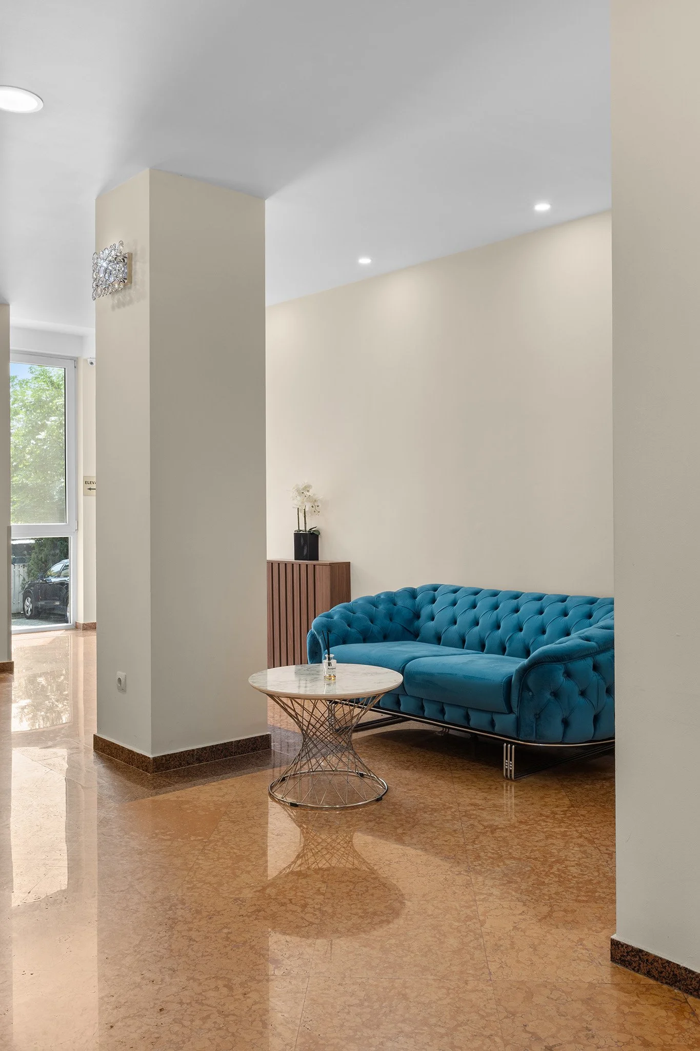 Interior of a modern living room with a bright blue tufted sofa, white walls, a marble-top side table, and a small wooden cabinet with white flowers, near an entrance with glass doors.