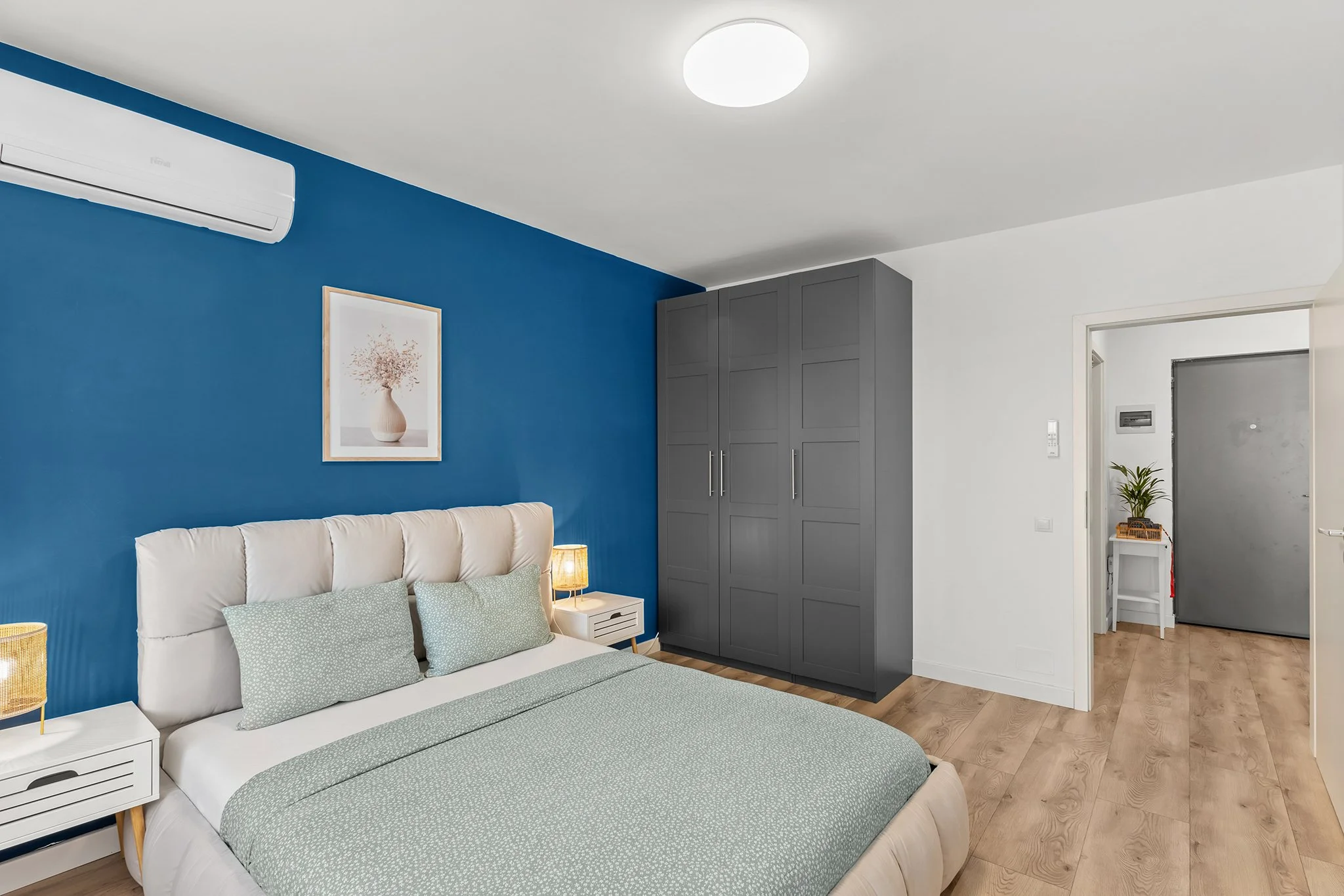 Modern bedroom with a bed covered in light green bedding, a padded headboard, blue accent wall, gray wardrobe, nightstands with lamps, potted plant, and wooden flooring.