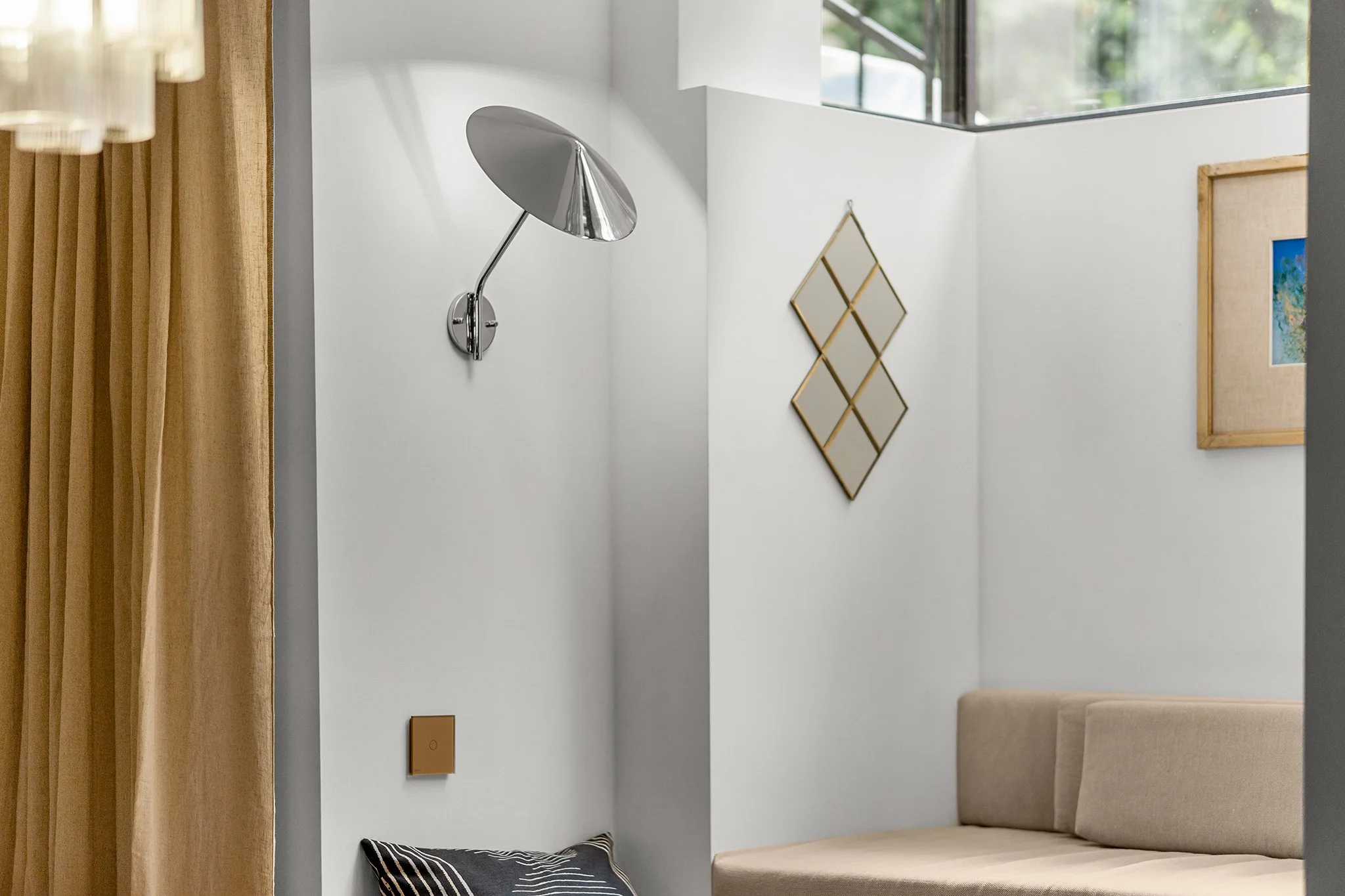 Interior view of a modern living space with a beige couch, wall decor including a gold-framed mirror and a art piece, a silver adjustable wall-mounted lamp, and a window with natural light.