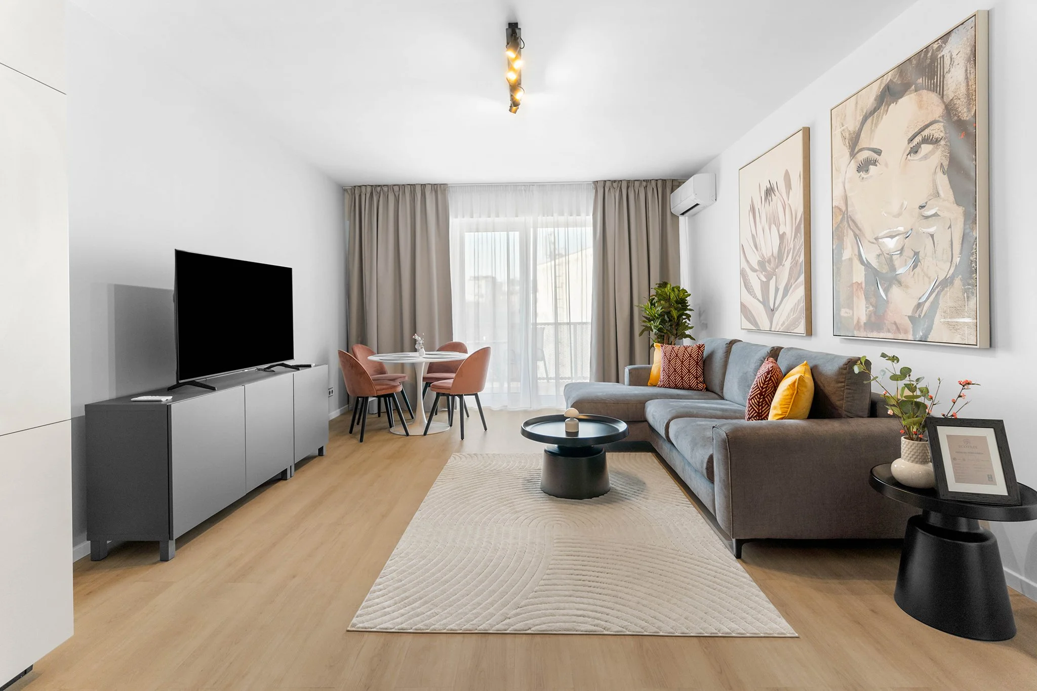 Modern living room with a gray sofa, colorful pillows, coffee table, black side table with a framed picture and vase, wall art, dining area, TV, beige curtains, hardwood floors, and a sliding glass door leading to balcony.
