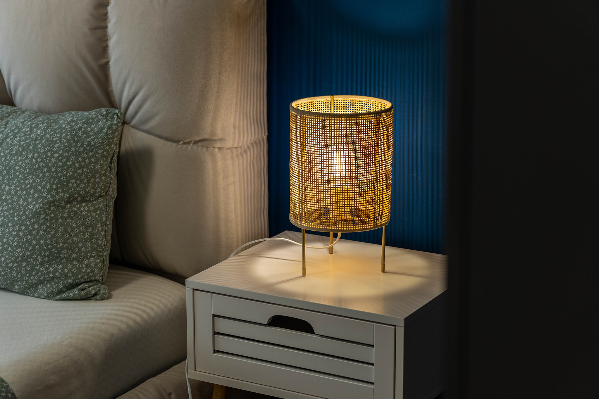 Bedroom scene with a yellow wicker lamp on a white nightstand, next to a beige upholstered headboard and a green floral pillow.