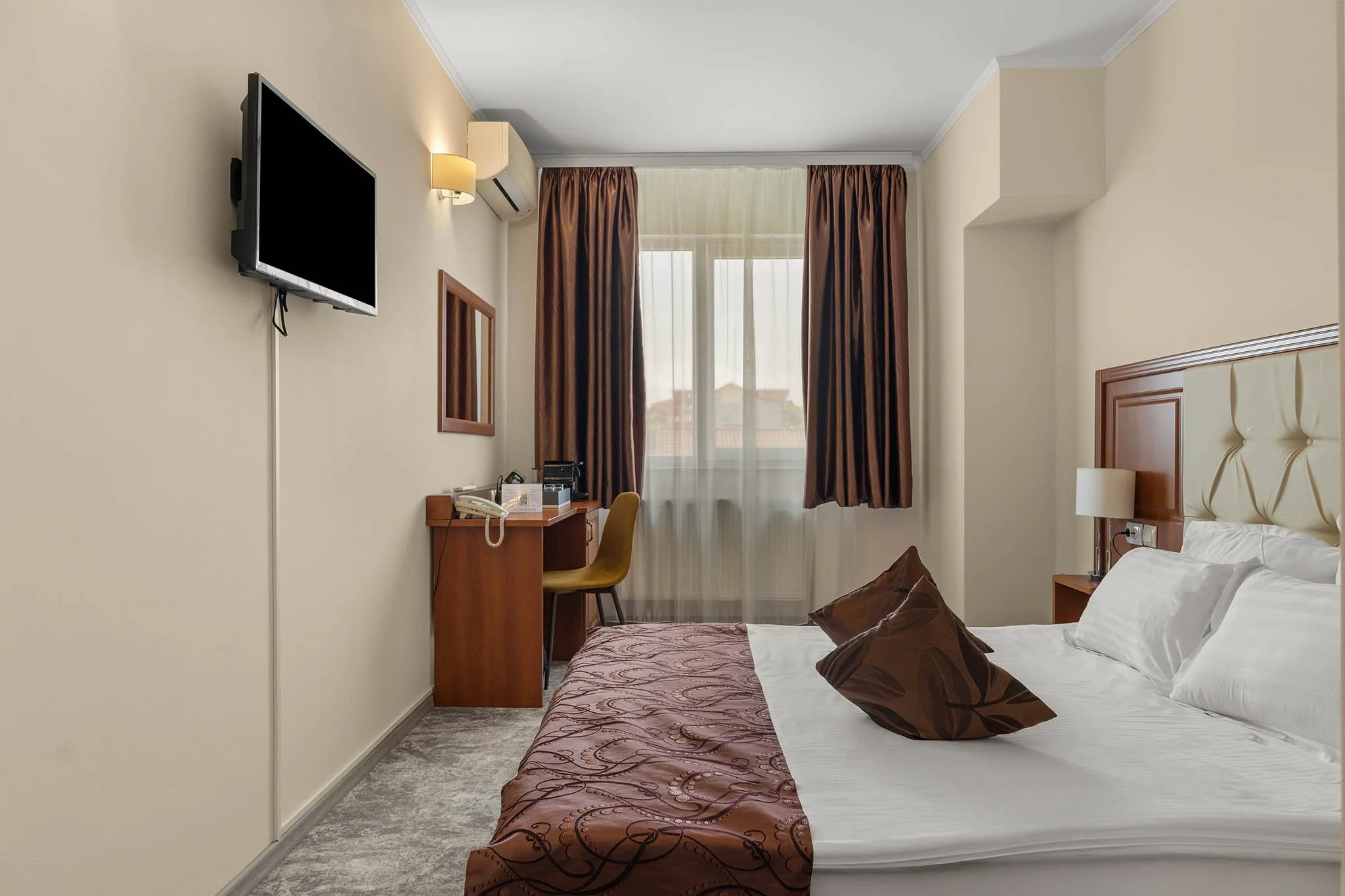 Hotel room with a bed, pillows, bedside lamp, window with brown curtains, desk with chair, wall-mounted TV, and air conditioner.