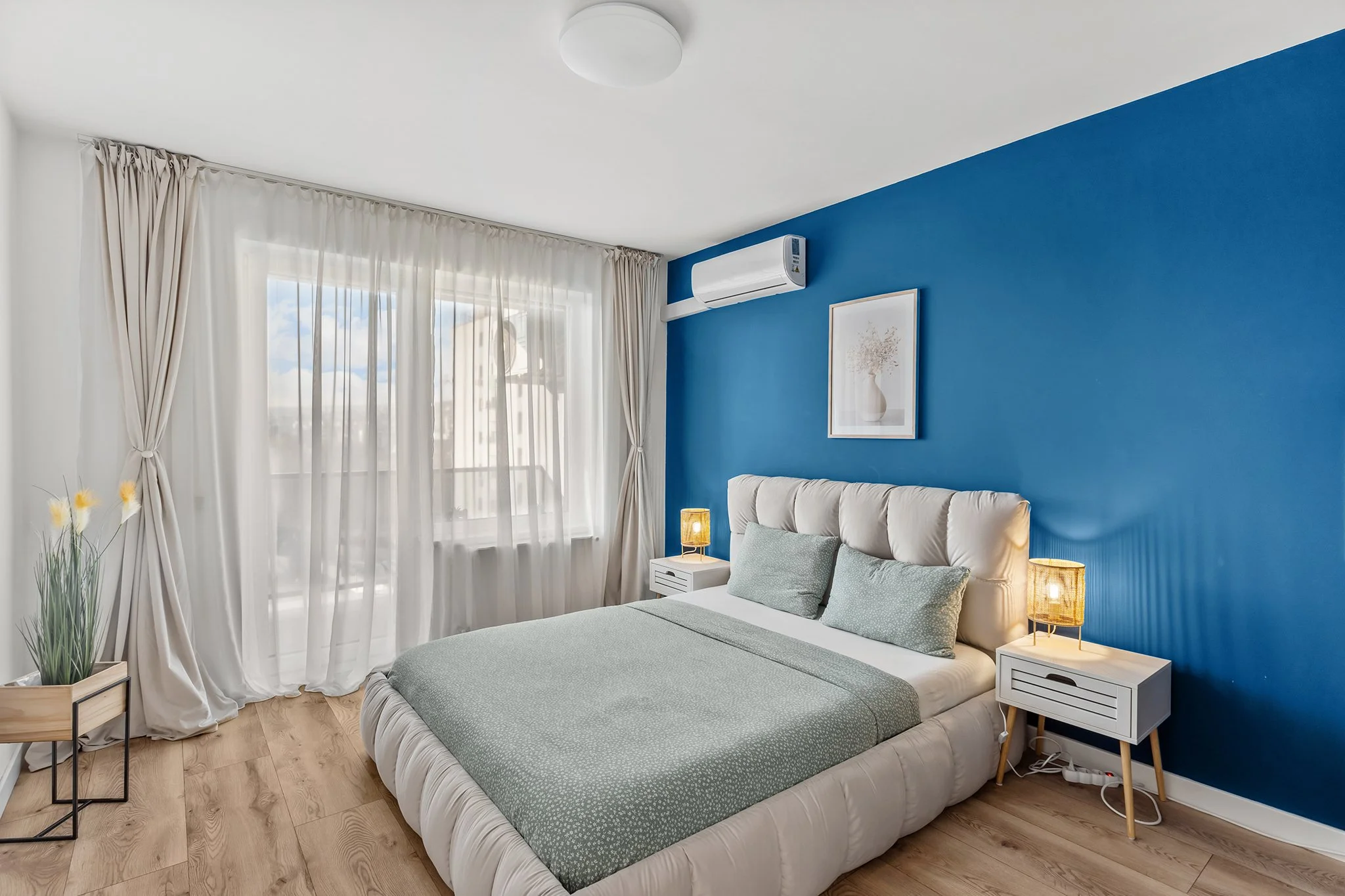Modern bedroom with a blue accent wall, white bed with green bedding and pillows, two white nightstands with yellow lamps, framed wall art, large window with sheer white curtains, air conditioning unit, and wooden flooring.