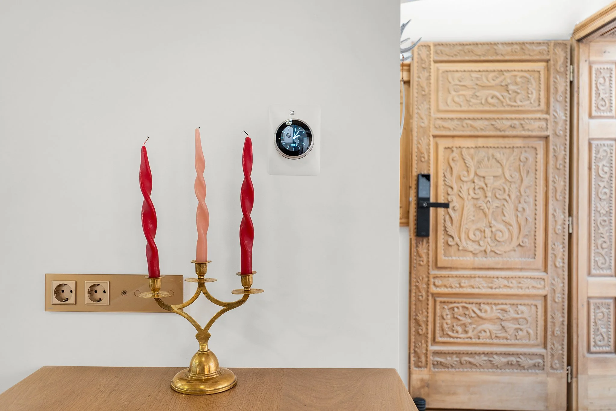 A gold candlestick holder with three twisted taper candles, two red and one pink, on a wooden table in front of a white wall. A digital thermostat is mounted on the wall, and part of an intricately carved wooden door is visible.