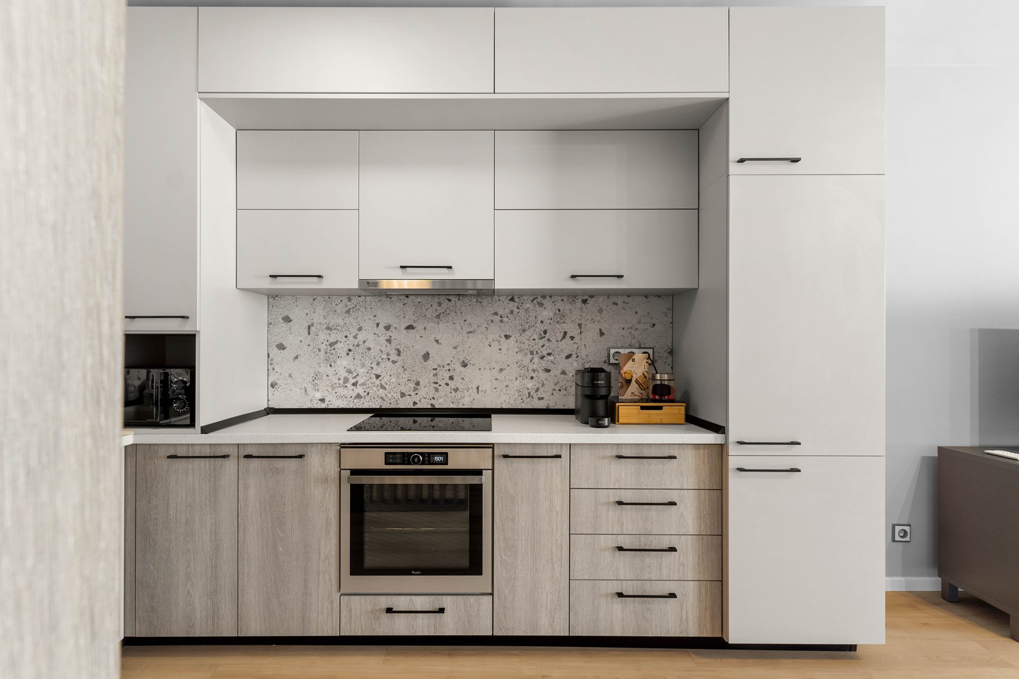 Modern kitchen with white and wood cabinets, a microwave, built-in oven, electric stovetop, coffee maker, and a terrazzo backsplash.