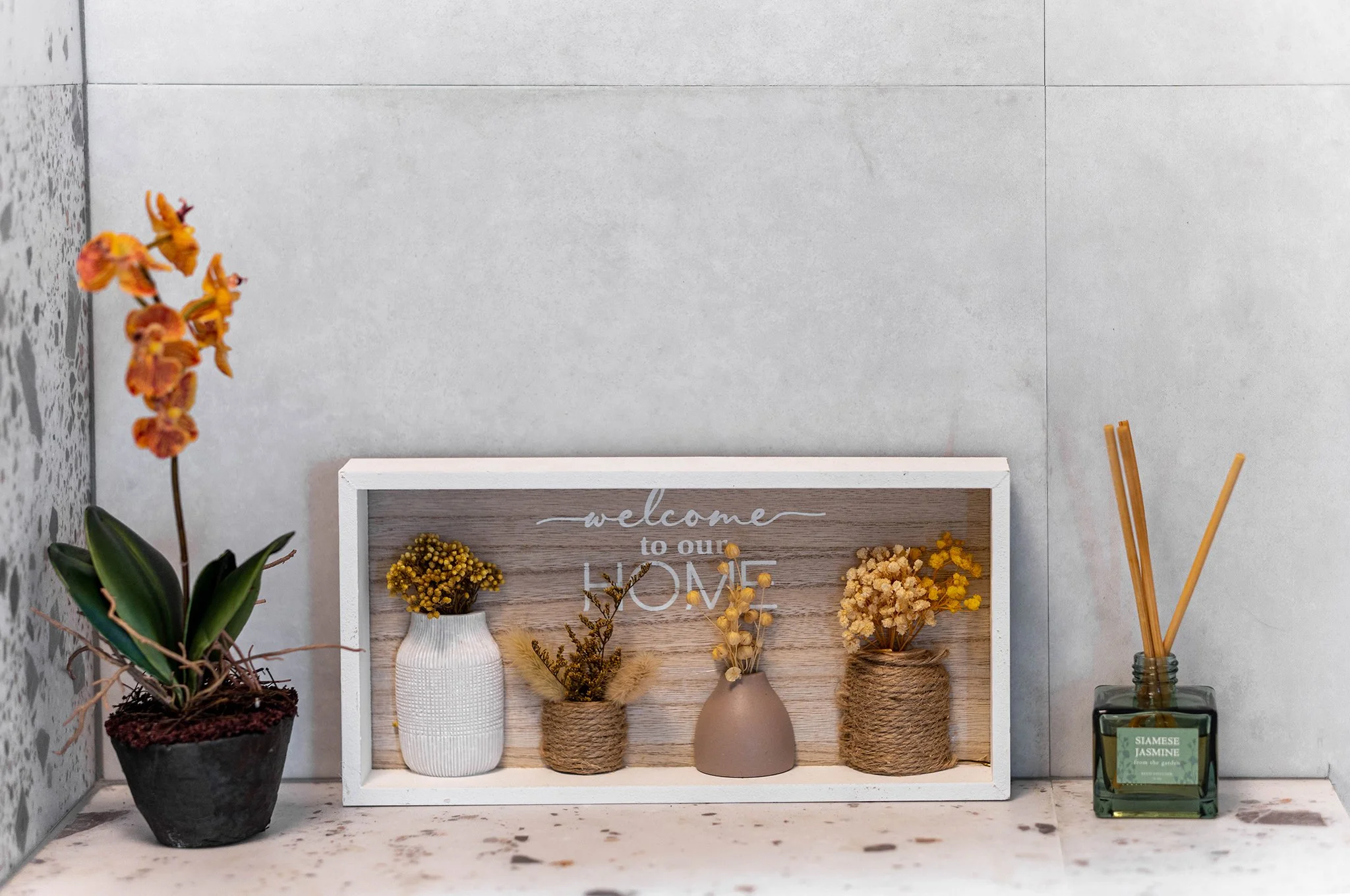Decorative setup with a potted orange orchid, a "Welcome to Our Home" sign featuring vases with dried flowers, and a Siamese Jasmine reed diffuser on a shelf.