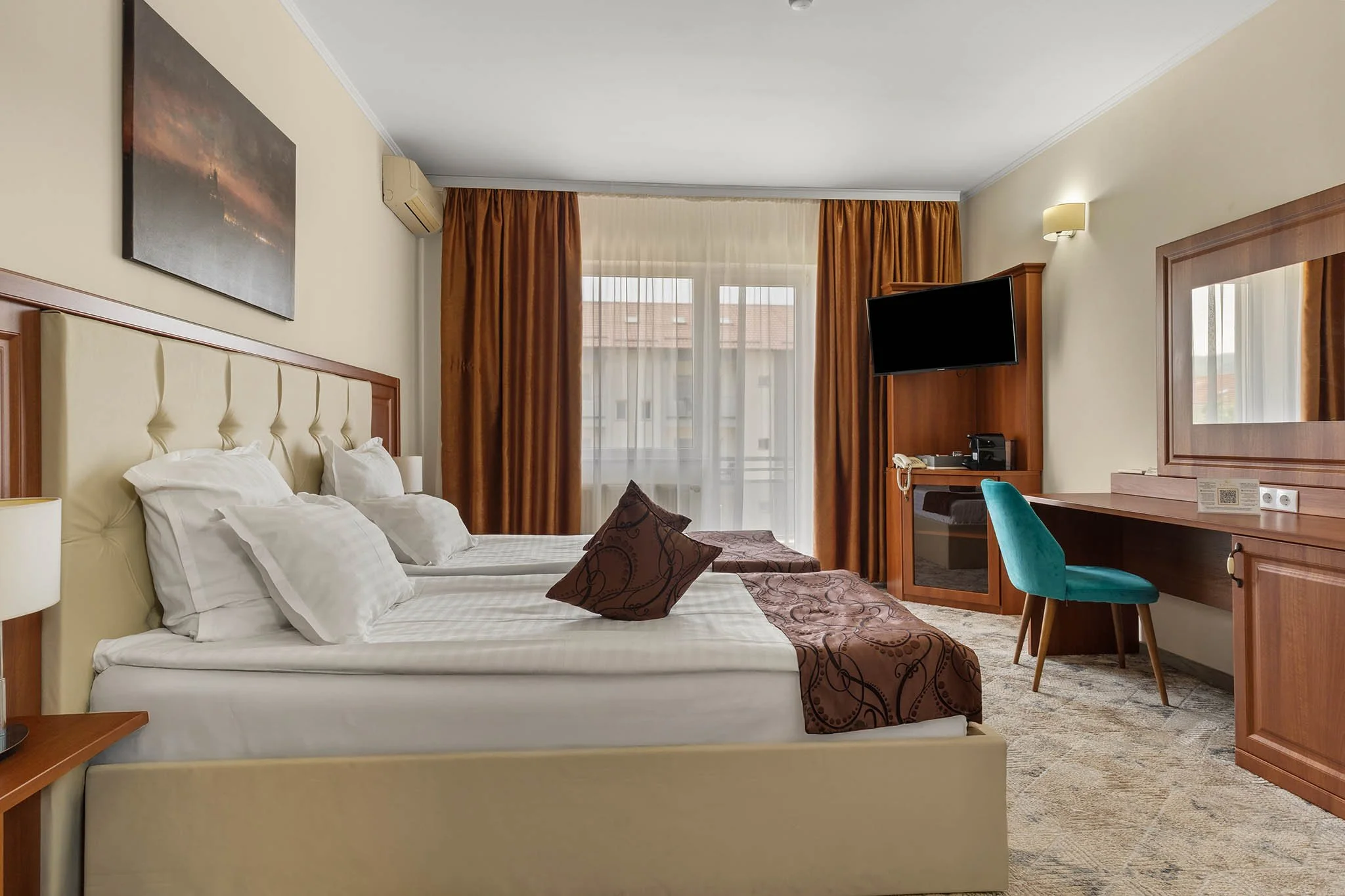 Hotel room with a bed, brown patterned bed runner, pillows, a desk with a blue chair, a wall-mounted TV, and a window with orange curtains.
