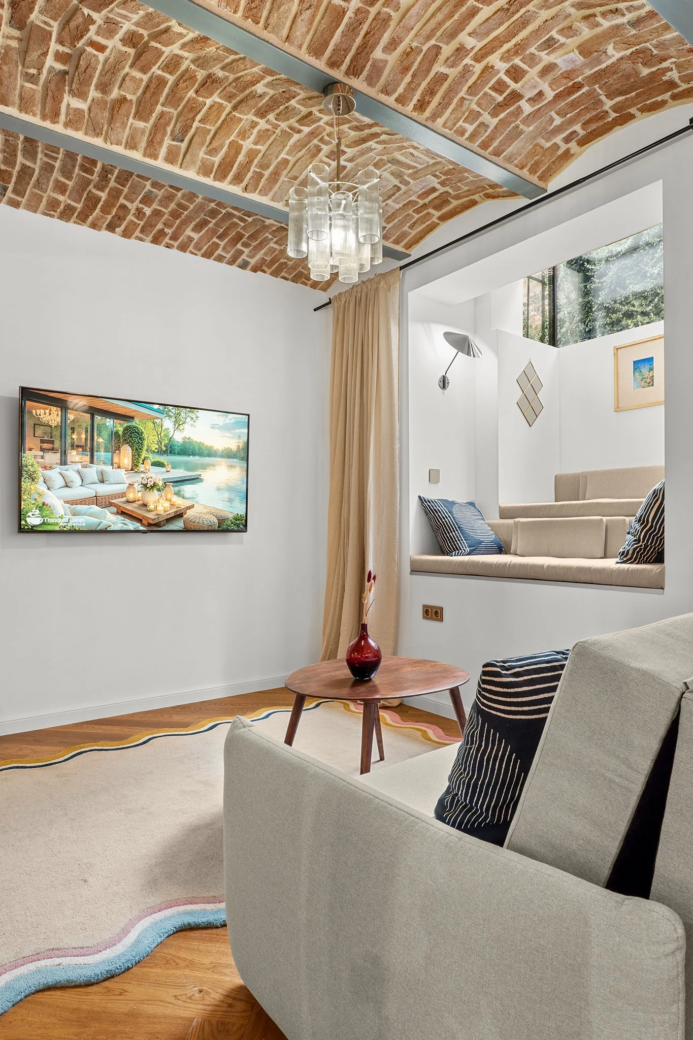 Living room with a brick vaulted ceiling, beige sofa with navy and white pillows, a wooden coffee table with a red vase, and a wall-mounted TV. There is a window seat with beige cushions and striped pillows, and a beige curtain partly drawn. The room