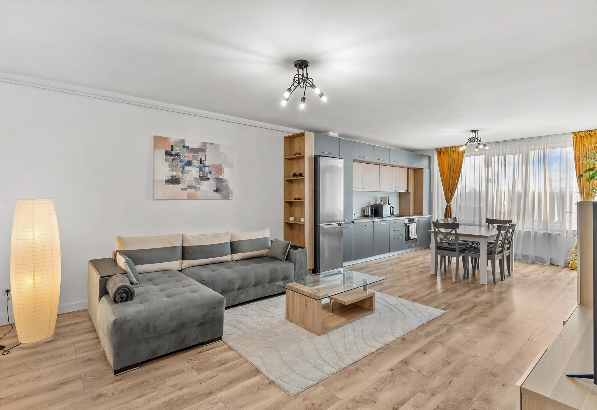 Modern open-concept living space with a gray sofa, wooden coffee table, contemporary kitchen with stainless steel appliances, dining area with four chairs, abstract wall art, and large window with yellow curtains.