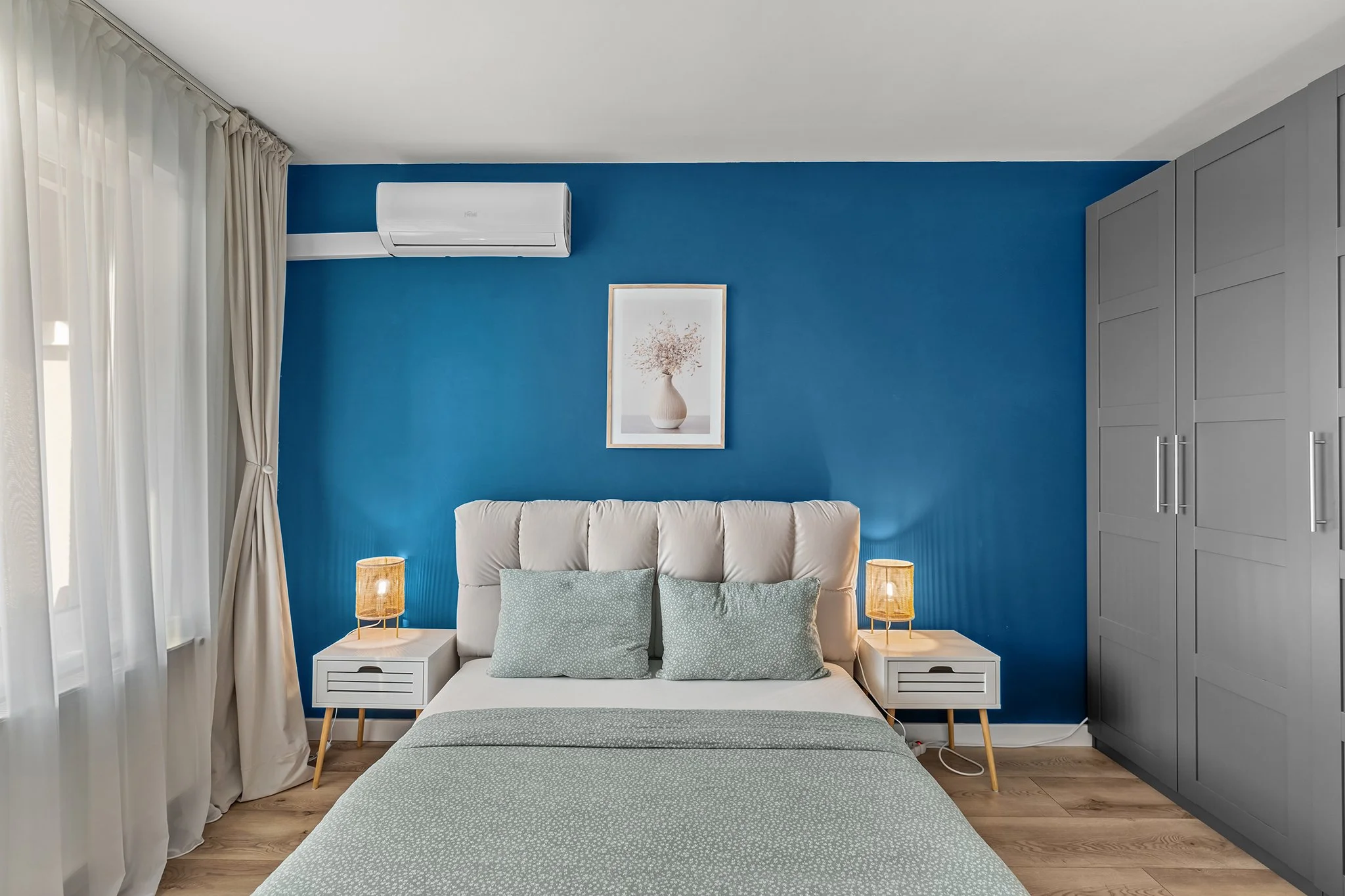 Modern bedroom with blue accent wall, white bed with patterned cushions, bedside tables with lamps, large gray wardrobe, and wall-mounted air conditioner.