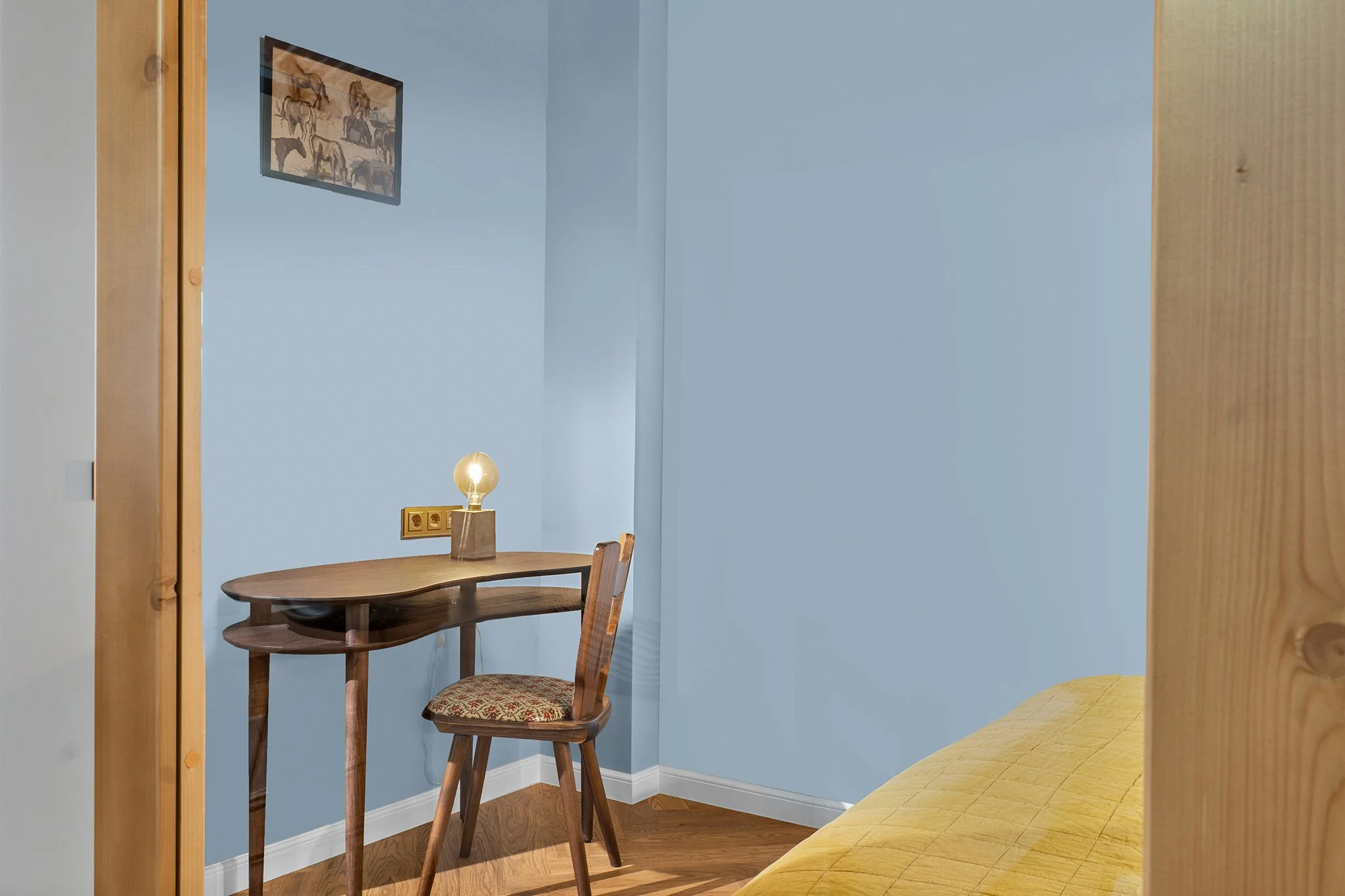 A small workspace corner with a vintage wooden desk, a matching chair with a patterned seat cushion, a yellow bed cover, a framed painting of horses on a blue wall, and a small table lamp with a glowing bulb.