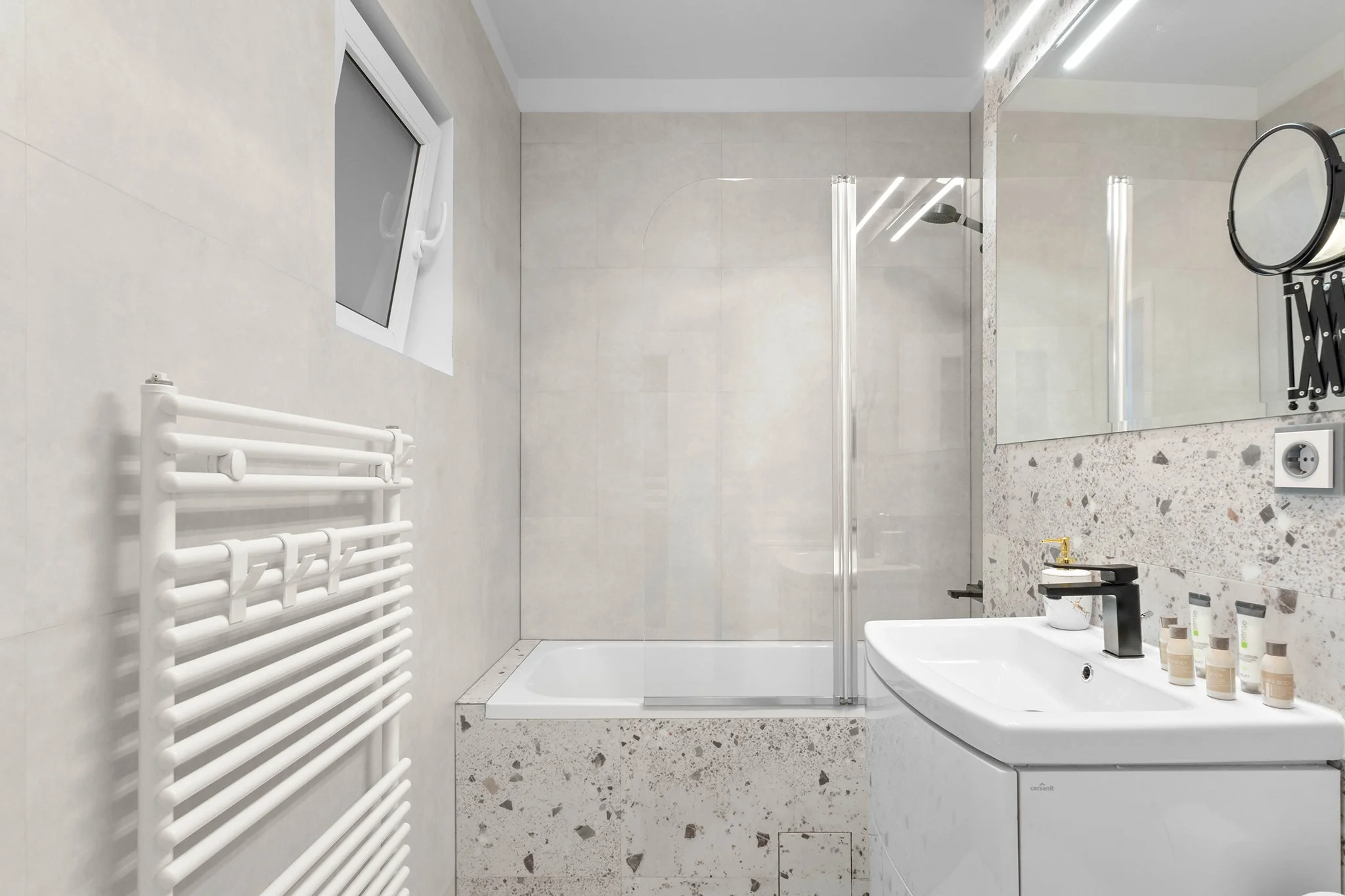 Modern bathroom with a bathtub and shower, white sink, terrazzo wall, towel radiator, and mirror with lights.