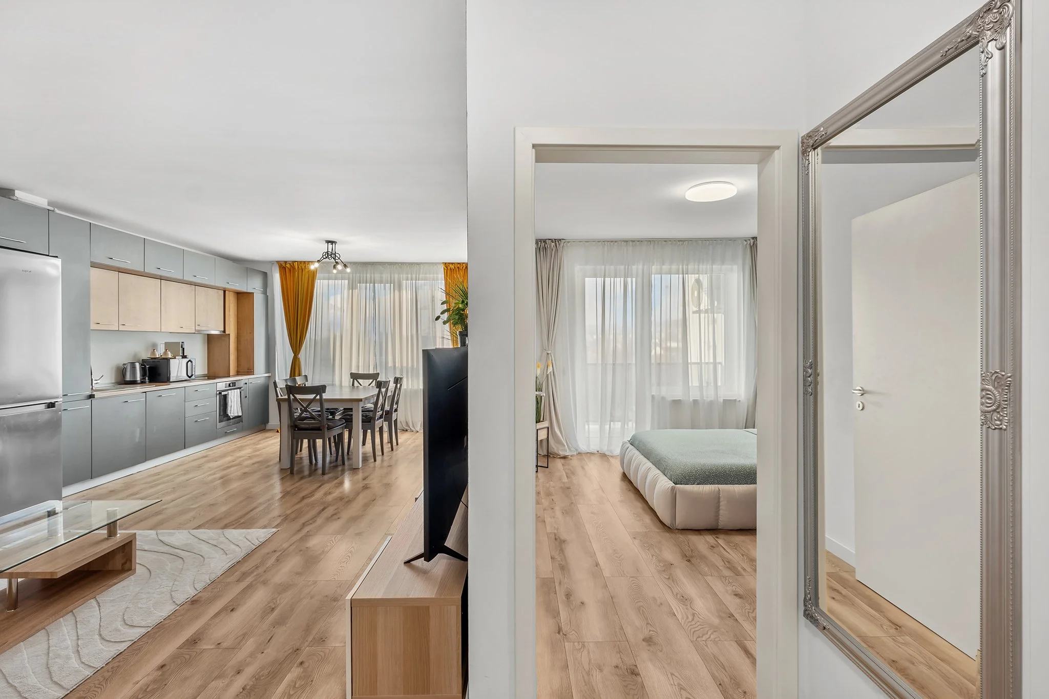 Modern open-plan living space with kitchen, dining area, and adjacent bedroom, featuring light wood floors, gray cabinetry, yellow curtains, and large windows.