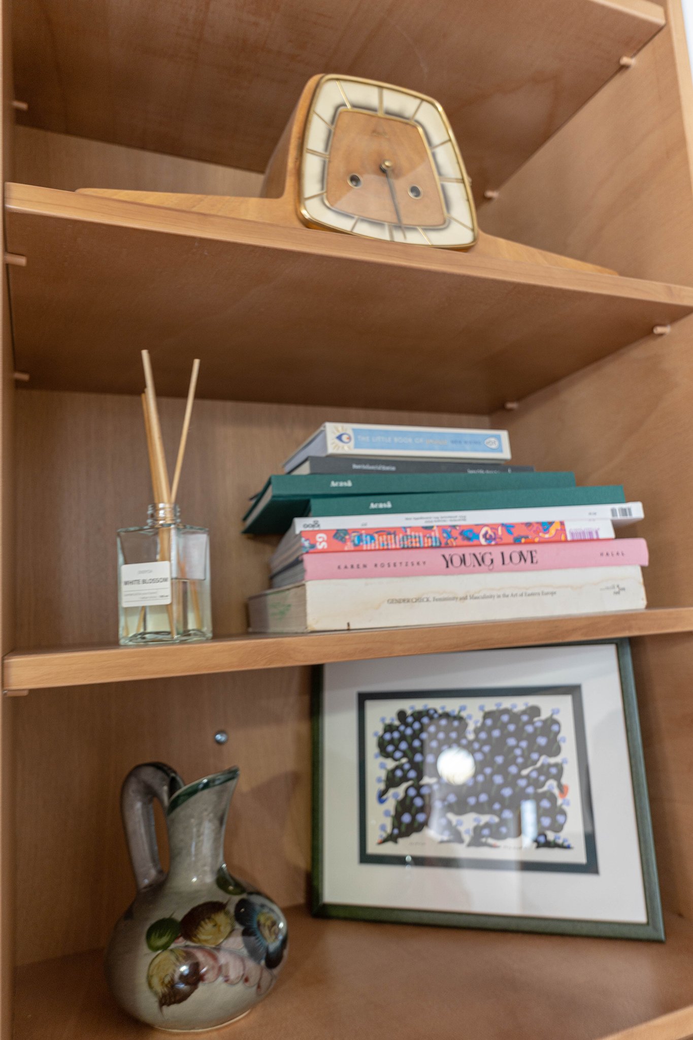 Wooden bookshelf with a vintage clock, books, a decorative pitcher, a framed picture, and a small jar with reed diffusers.