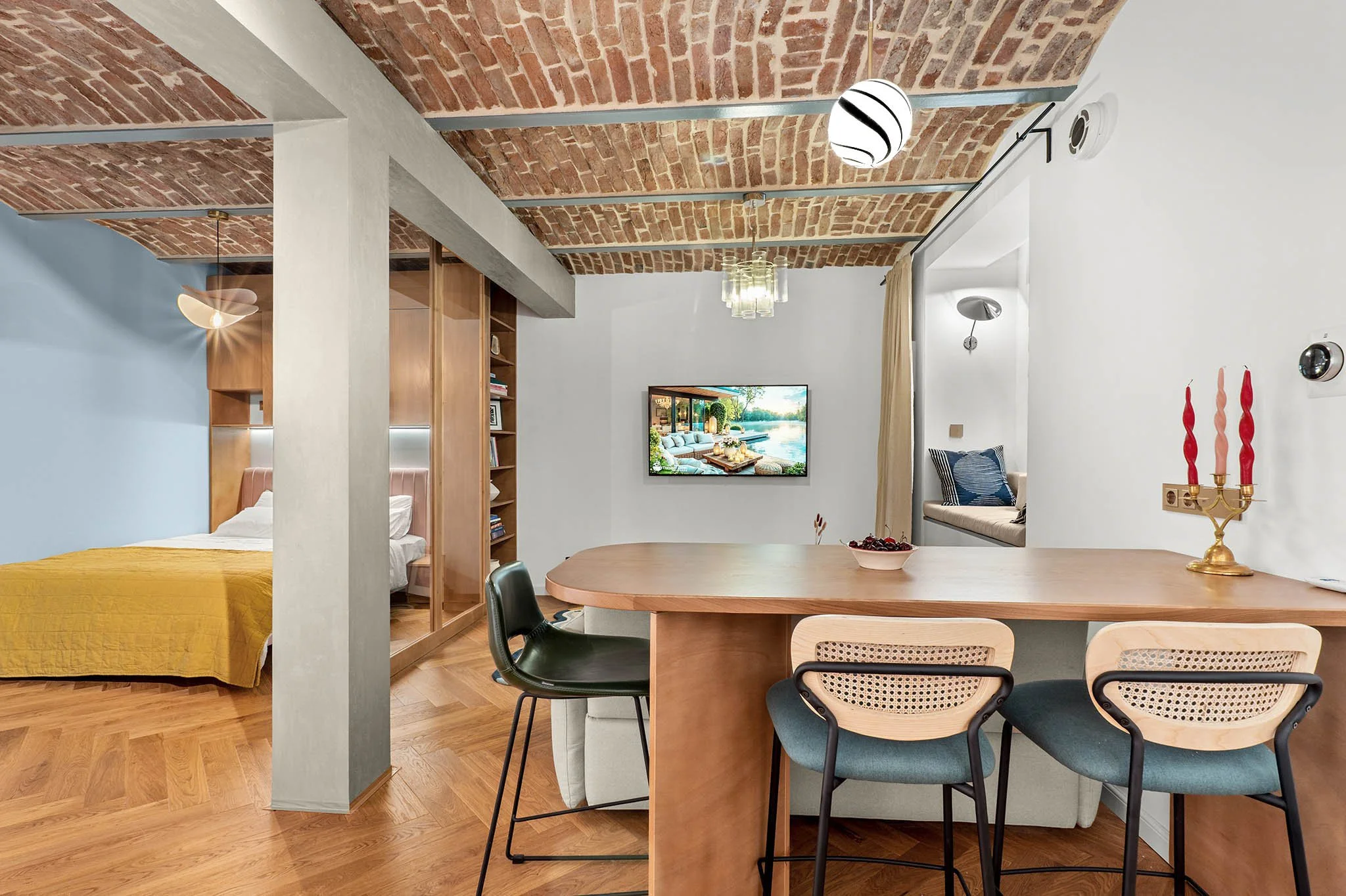 Open-concept living space with a bedroom, dining area, and sitting nook. Exposed brick ceiling, wooden flooring, a wall-mounted TV, and modern lighting fixtures. The space includes a yellow bedspread, a TV, chairs around a wooden table, and a cozy no