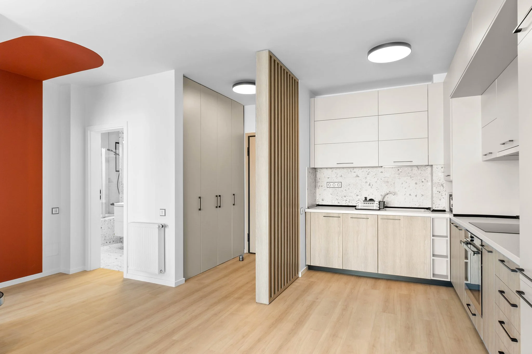Modern kitchen interior with wooden cabinets, white countertops, and a light-colored parquet floor; features a red accent wall and vertical wooden slats.