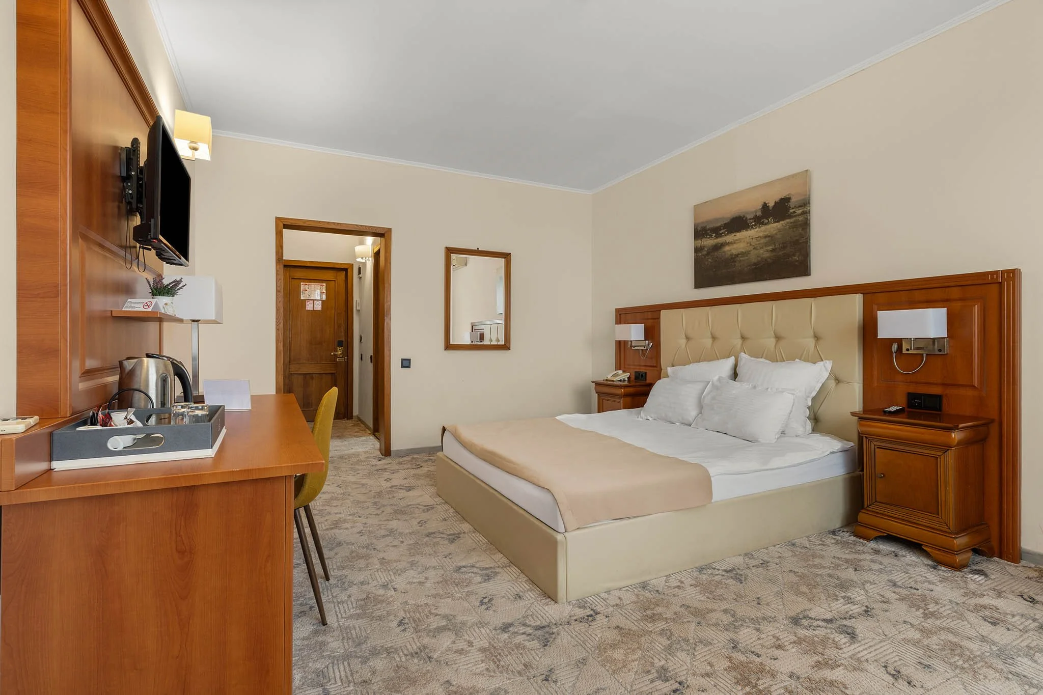 Hotel room with a bed, bedside tables, wall lamps, a mirror, a TV, and a desk with an electric kettle and chairs.