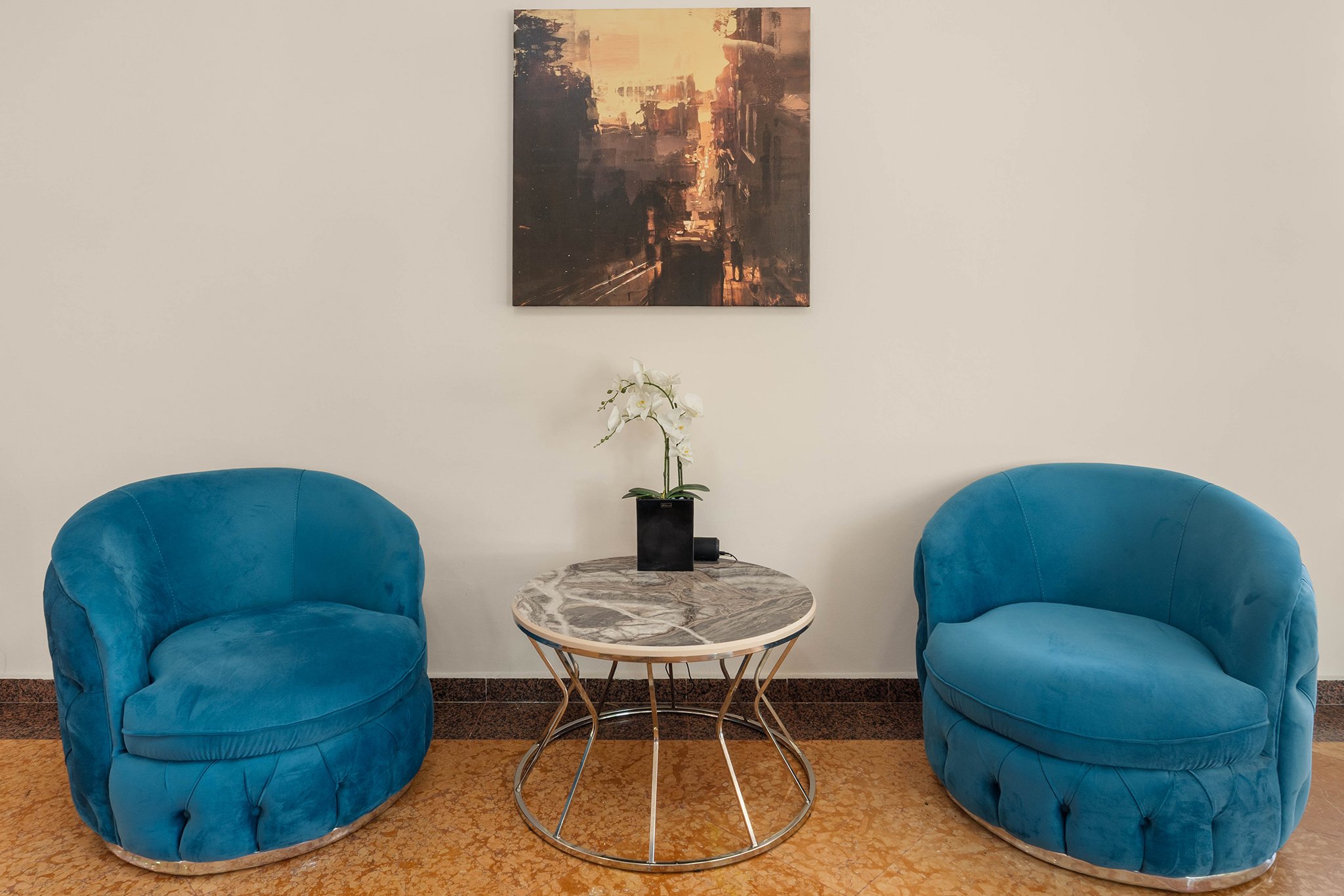 Two blue velvet armchairs are situated on either side of a round marble-top table with a orchid plant in a black pot, against a white wall with a cityscape painting hanging above.