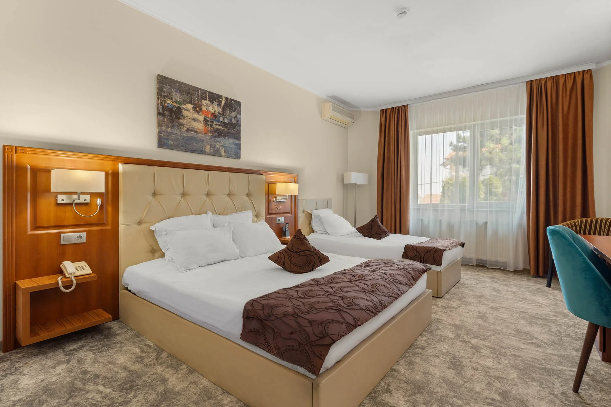 Hotel room with two beds, a large window with orange curtains, a painting above the headboards, and a small table with a teal chair.