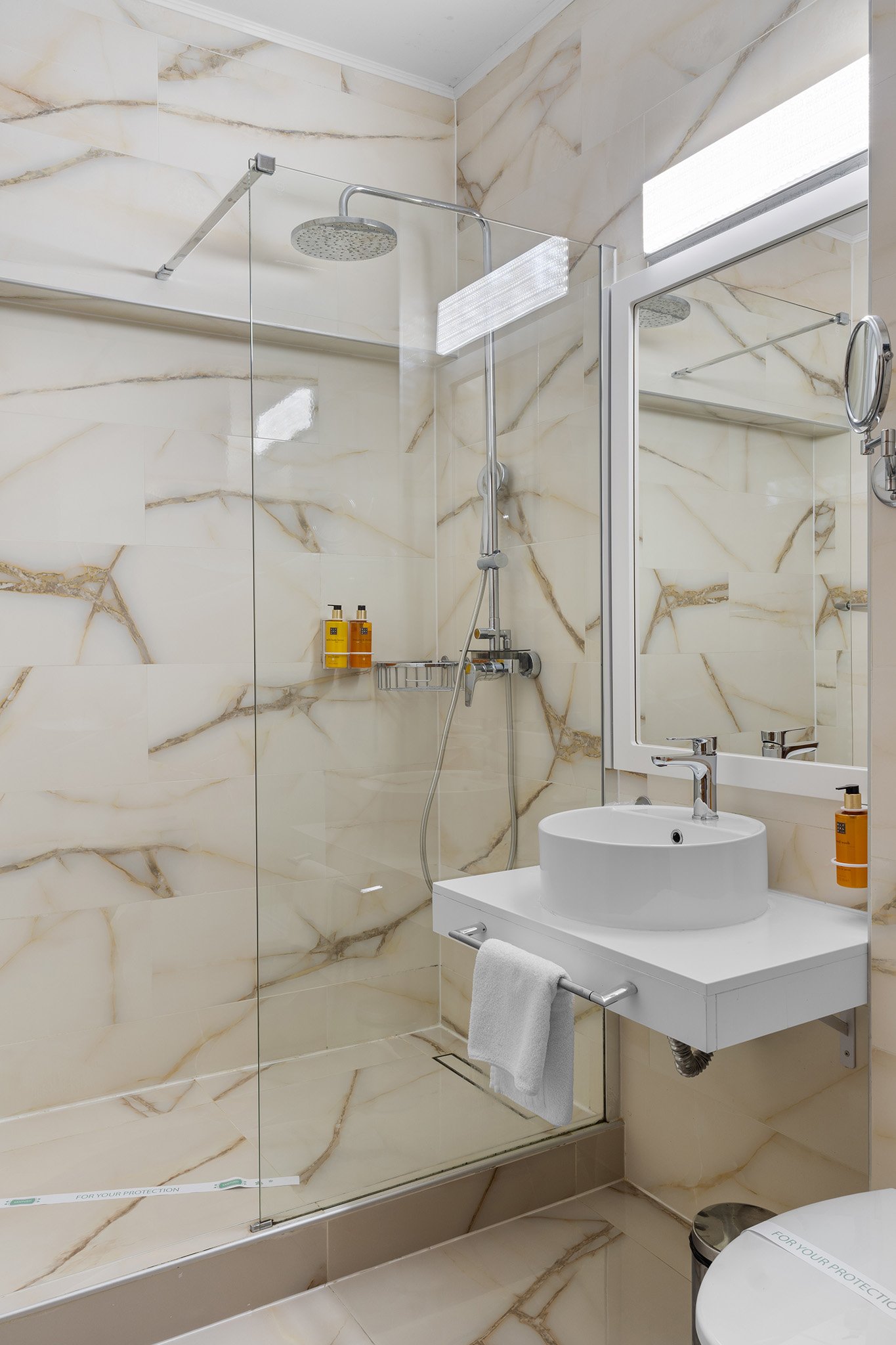 Modern bathroom with marble tiles, glass shower enclosure, wall-mounted sink with mirror, and vanity with towel, along with toiletries and a window.