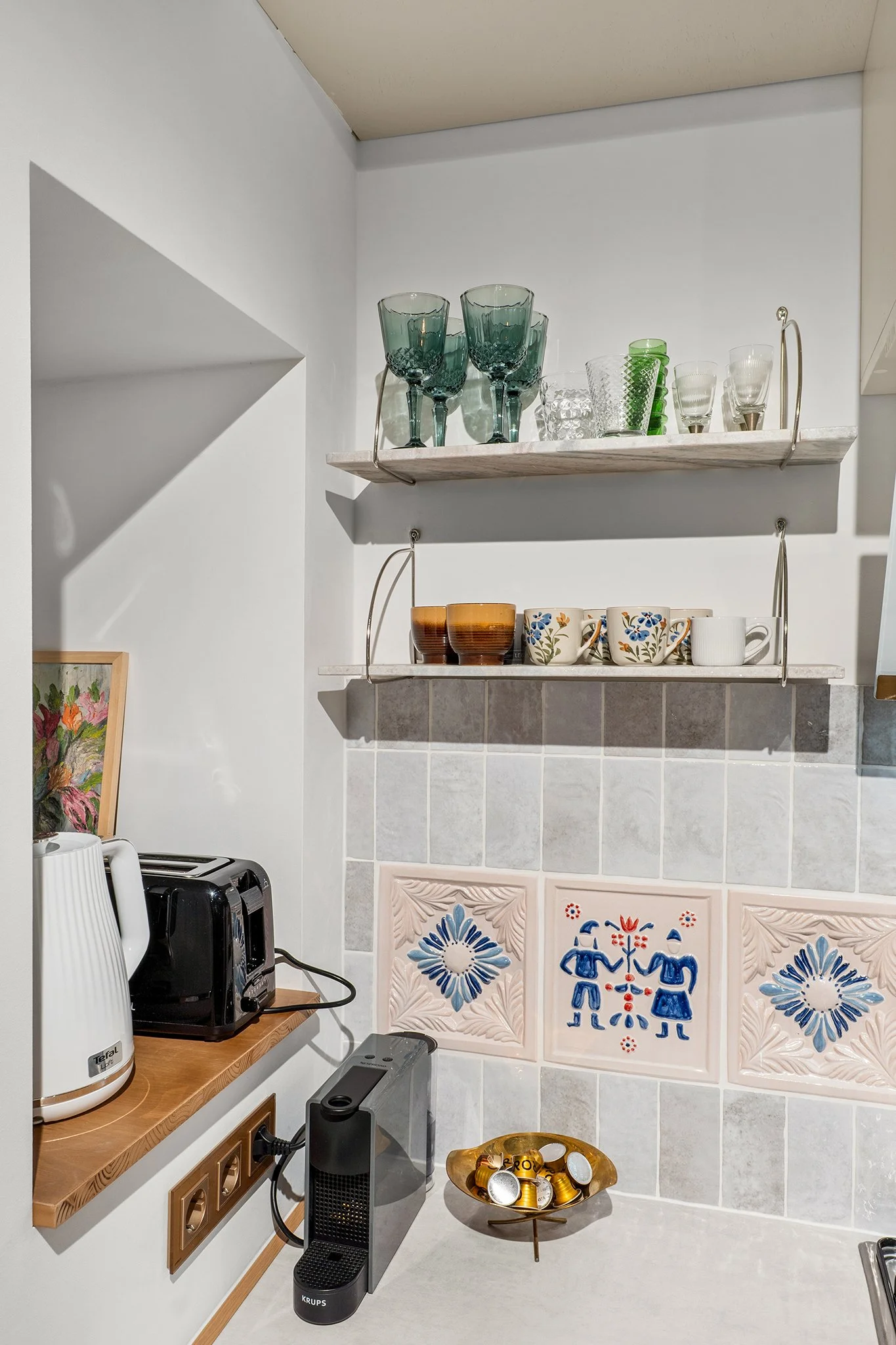 Kitchen corner with glassware on wall shelves, coffee machines, a Koenigsegg pod, and decorative tiles featuring traditional blue and pink patterns.