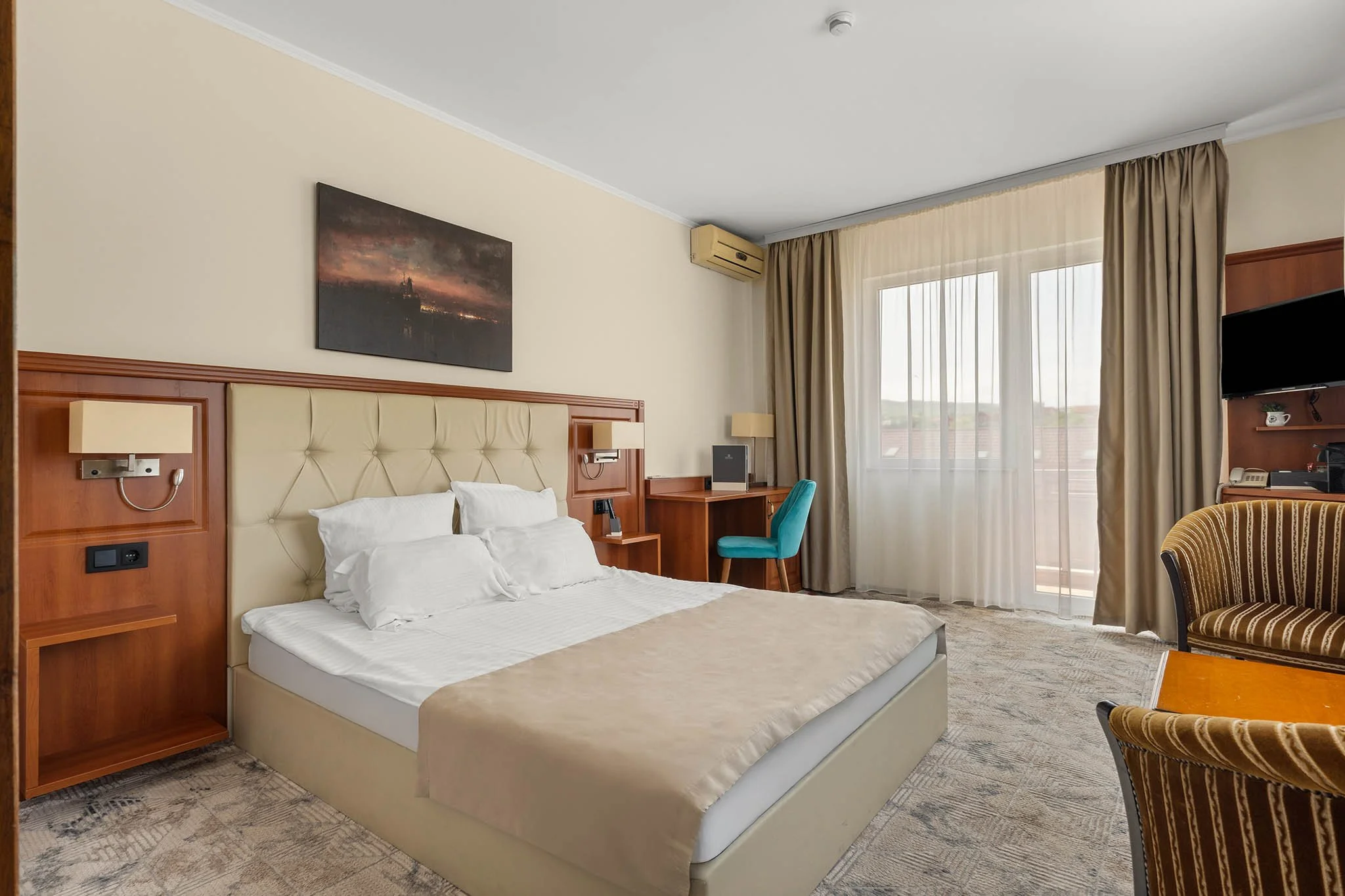 Hotel room with a queen bed, beige headboard, wall-mounted lights, desk with a turquoise chair, and large window with beige curtains.