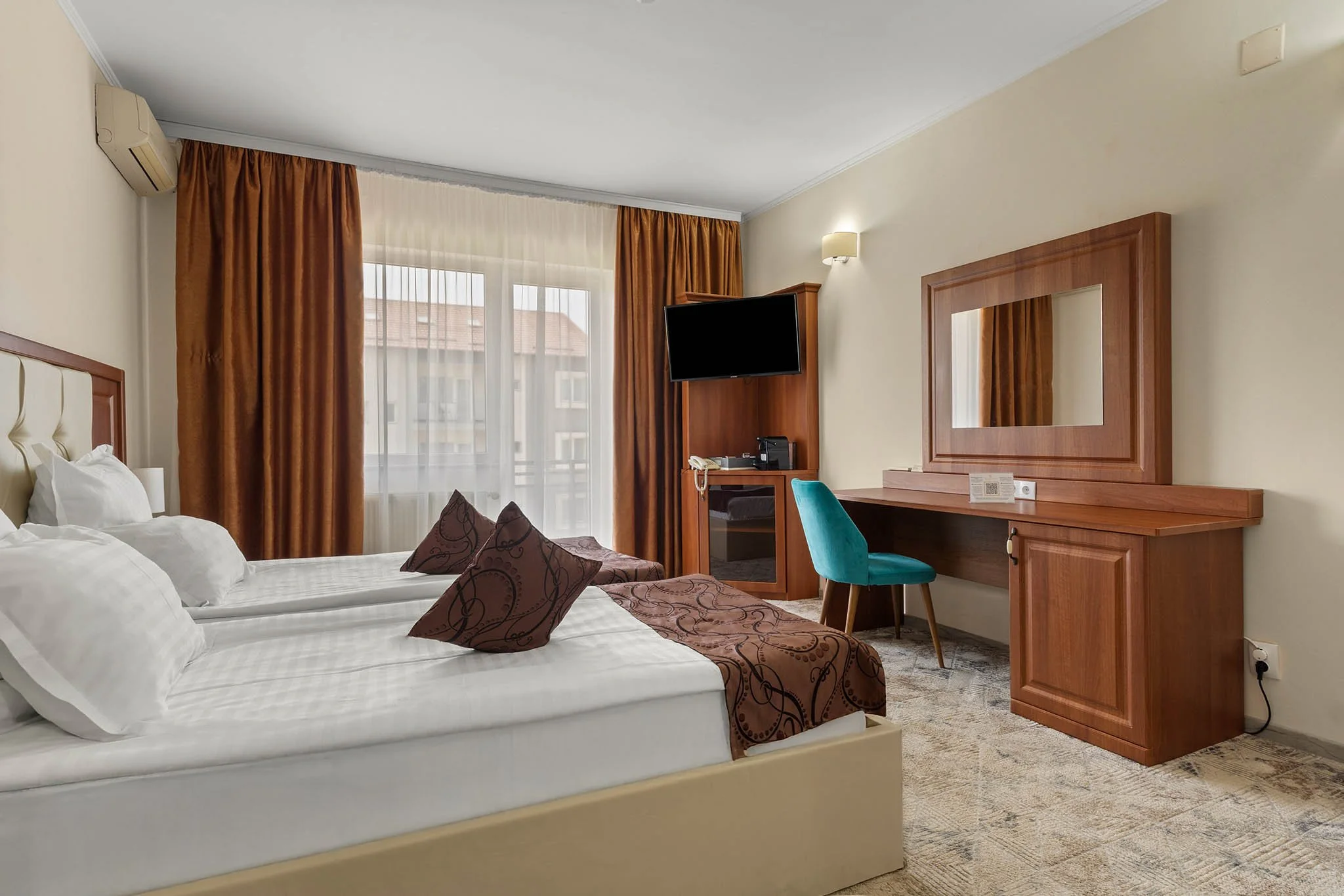 Hotel room with large bed, brown pillows, brown blanket, window with curtains, mounted TV, desk with mirror, teal chair, and carpeted floor.