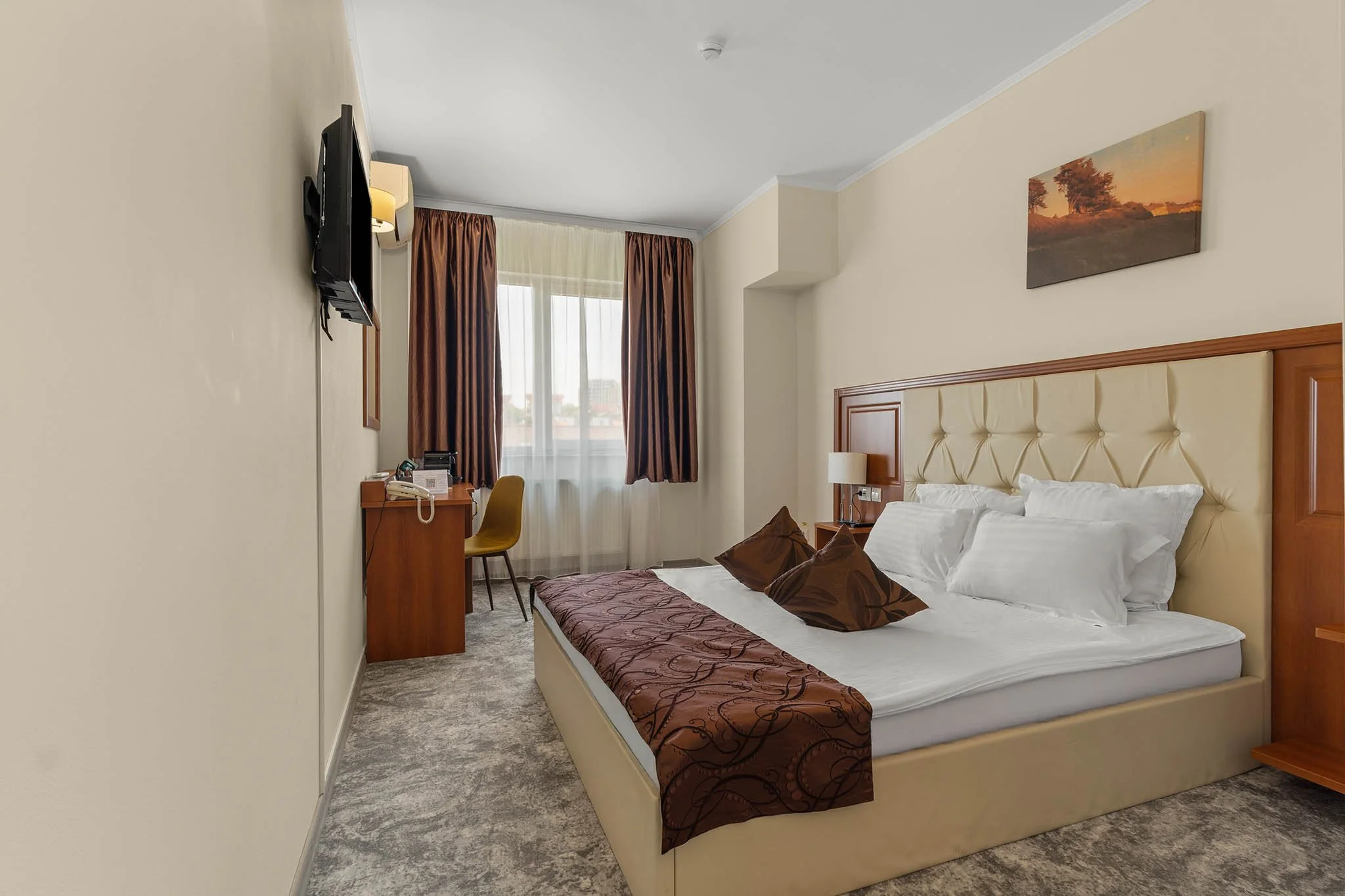 Hotel room with a large bed, wooden headboard, brown decorative pillows, wall-mounted TV, window with brown curtains, and a small desk with a yellow chair.