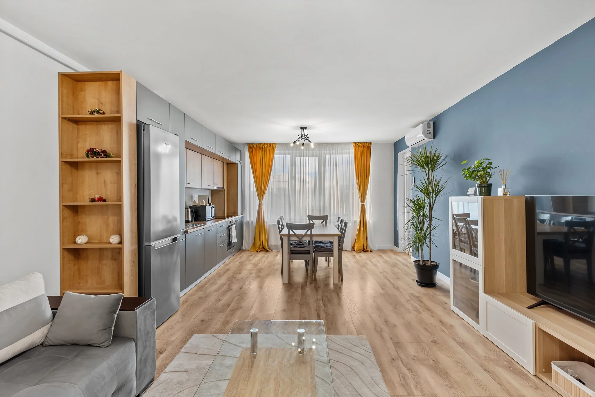 Open-concept living room and kitchen with wooden flooring, gray and white furniture, potted plants, and yellow curtains.