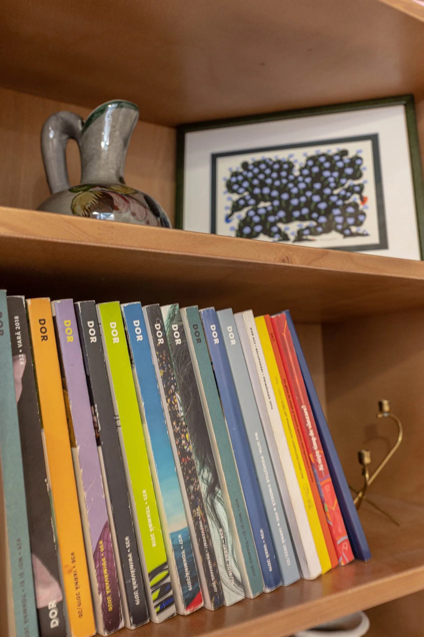 A wooden bookshelf containing colorful comic books, a decorative jug with flowers, a framed artwork of a dark tree with light purple leaves, and a small brass lamp with an adjustable arm.