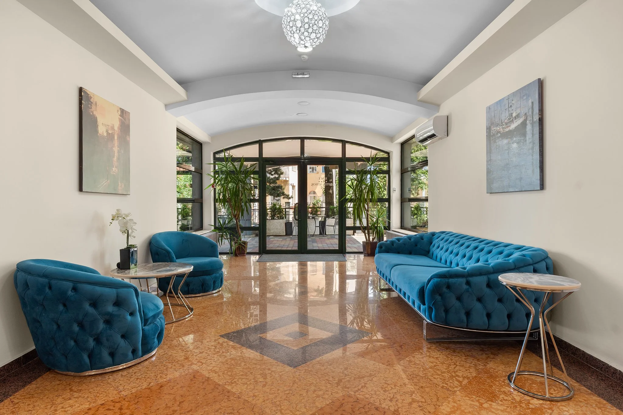A modern lobby with two blue velvet sofas, two blue velvet armchairs, small round tables, potted plants, and large windows, with a glass door entrance.