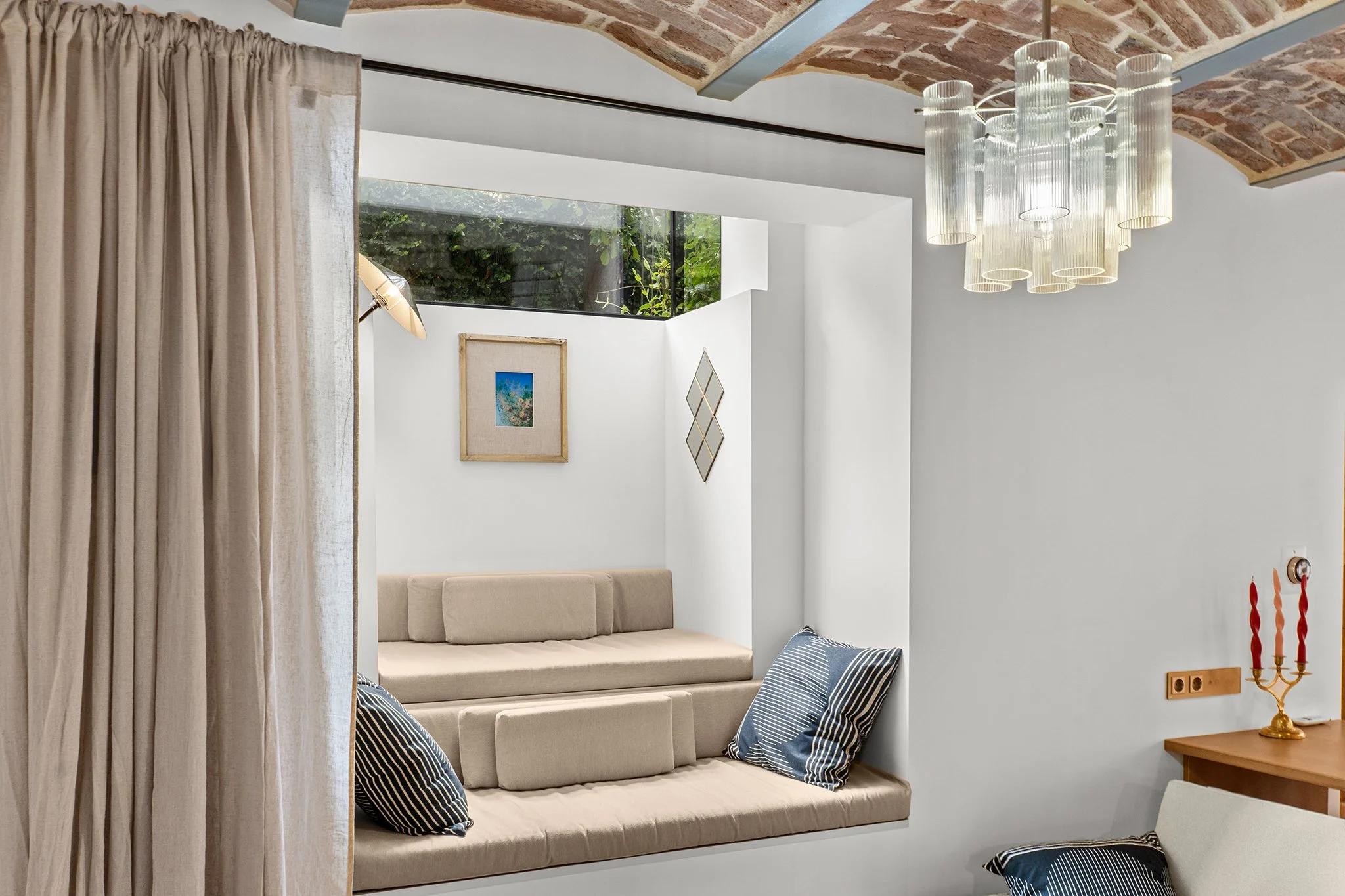 Cozy window nook with beige cushioned seating, surrounded by white walls, blue-striped pillows, framed artwork, a chandelier, and a wooden table with a gold candle holder and candles.