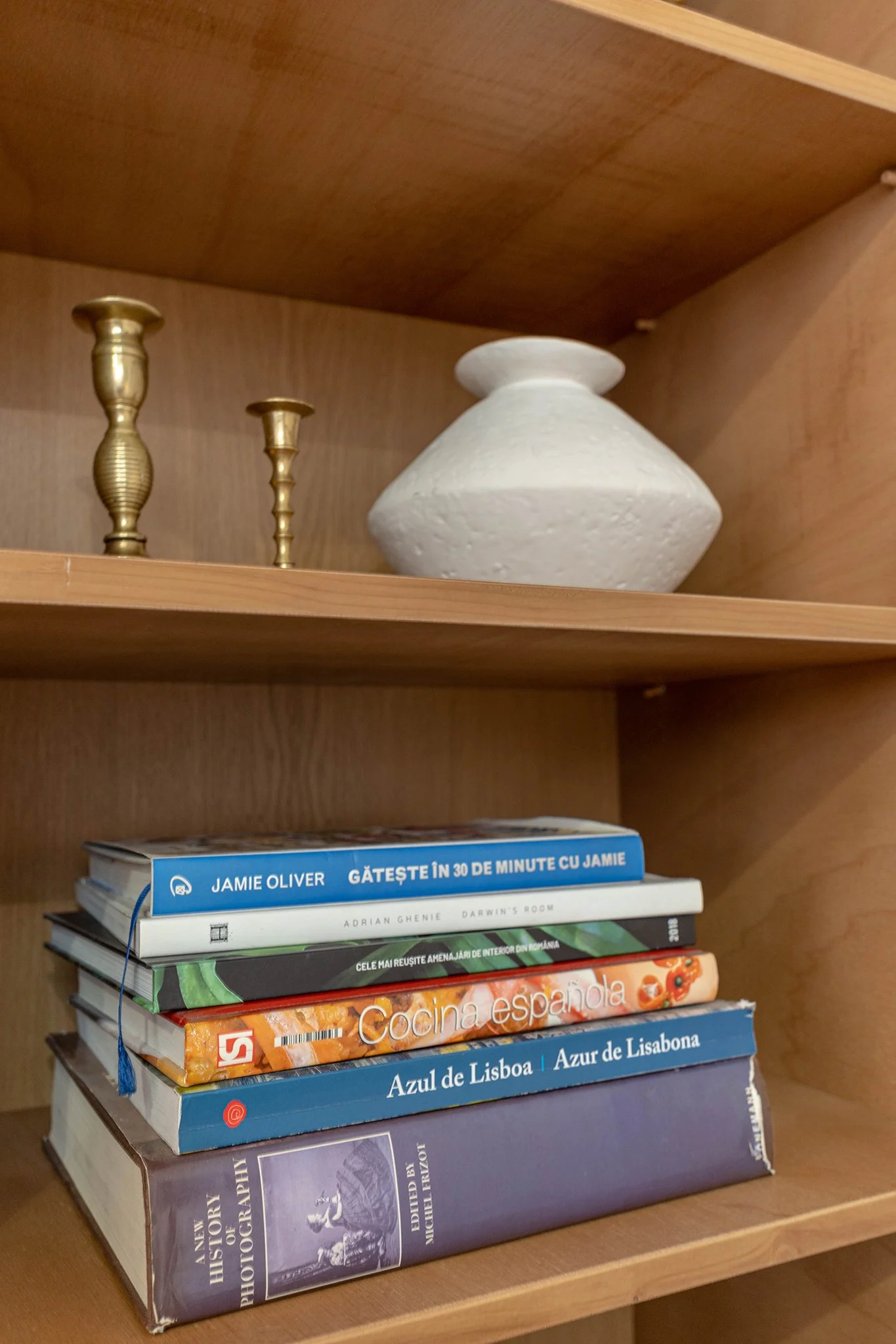 A bookshelf with two brass candlesticks, a white decorative vase, and a stack of books including titles about Jamie Oliver, interior design, Spanish cuisine, and Lisbon, Portugal.