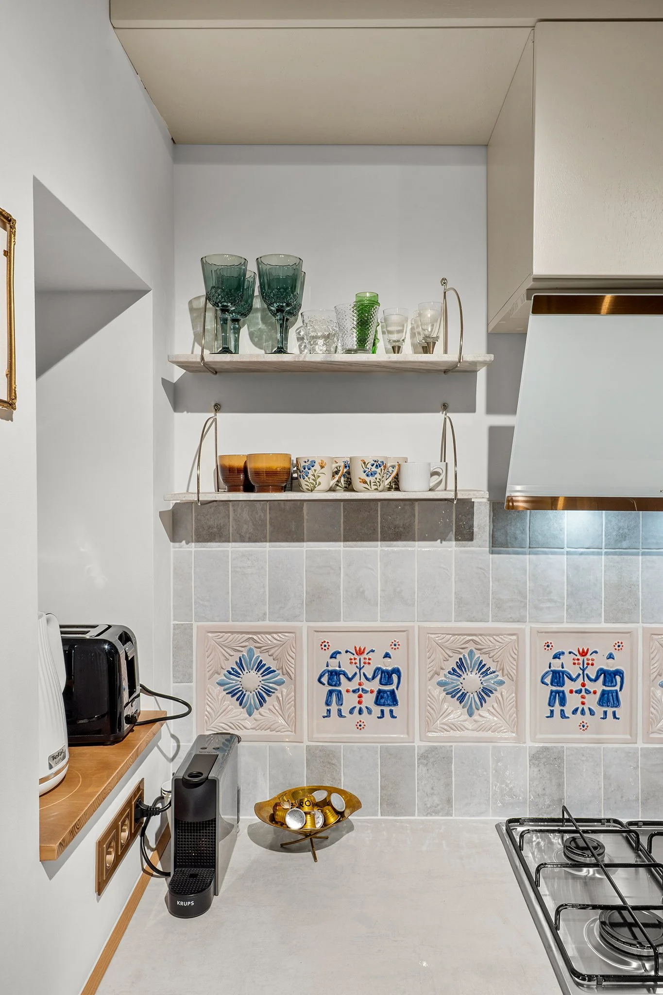 Kitchen with open shelves holding glassware and cups, tiled backsplash with decorative blue, pink, and red tiles, black coffee machine, toaster, and a bowl of coffee capsules on the countertop.
