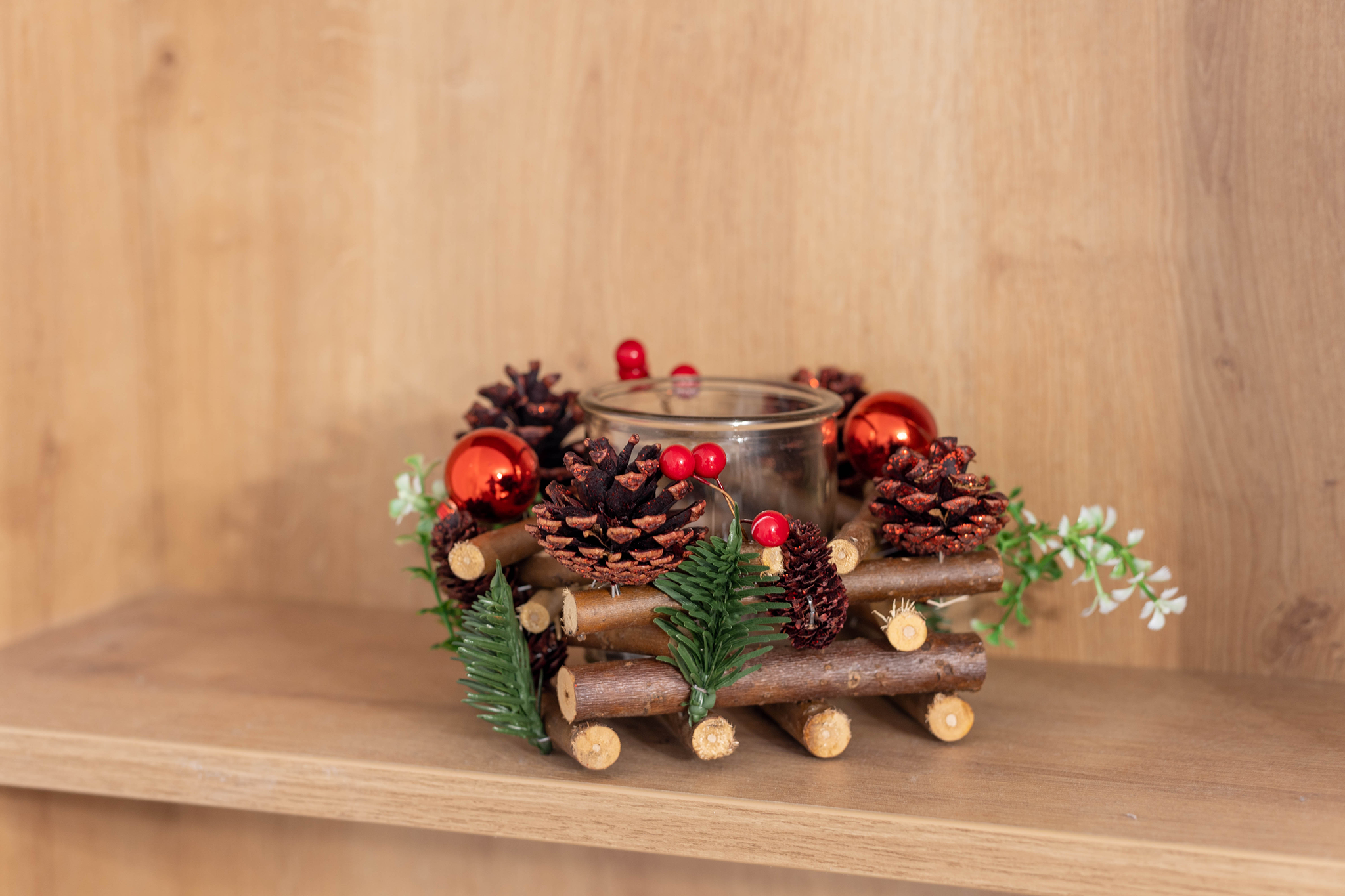 Festive candle holder decorated with pinecones, red berries, and evergreen leaves on a wooden shelf.