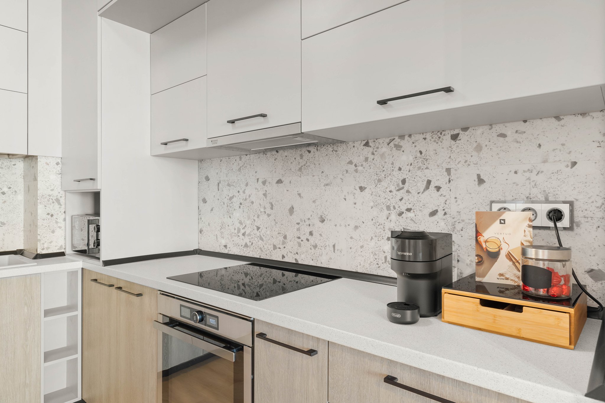 Modern kitchen with white cabinets, speckled backsplash, an induction cooktop, Nespresso coffee machine, and a wooden box holding coffee accessories.