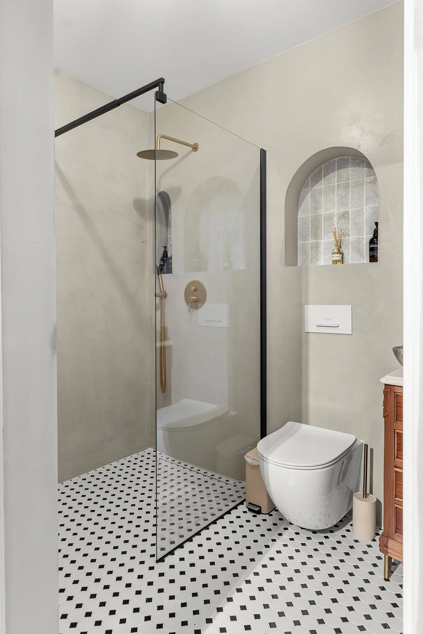Modern bathroom with a walk-in shower, glass partition, beige walls, black and white patterned tile floor, wall-mounted toilet, small beige trash bin, wooden vanity, and decorative niches with toiletries.