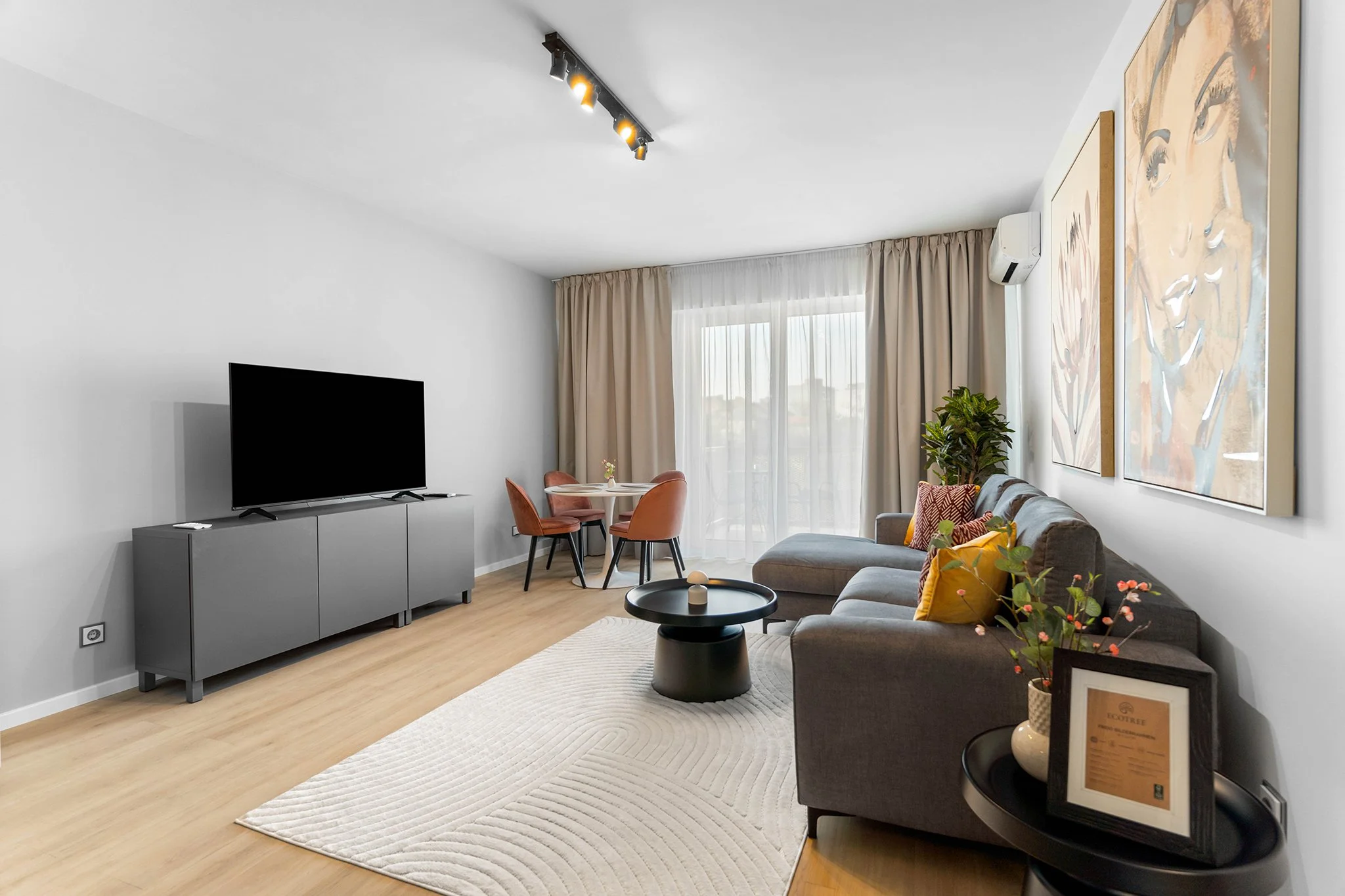 Living room with gray sofa, black coffee table, TV on gray cabinet, round dining table with 3 rose-colored chairs, large window with beige curtains, and wall art.