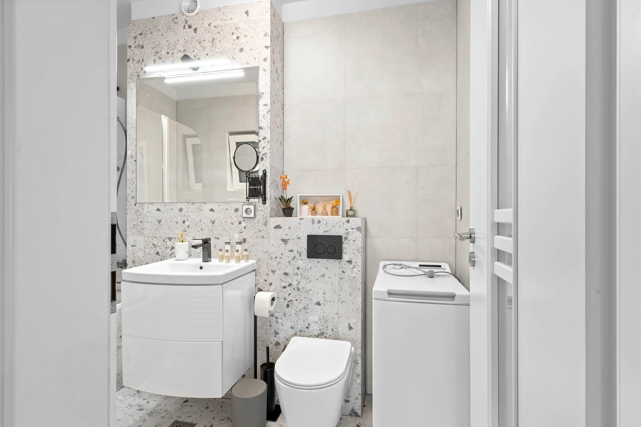 Modern bathroom with a white vanity, large mirror, toilet, and washing machine. Terrazzo wall and neutral tiles enhance the sleek design. Small potted plants and toiletries add decorative touches.