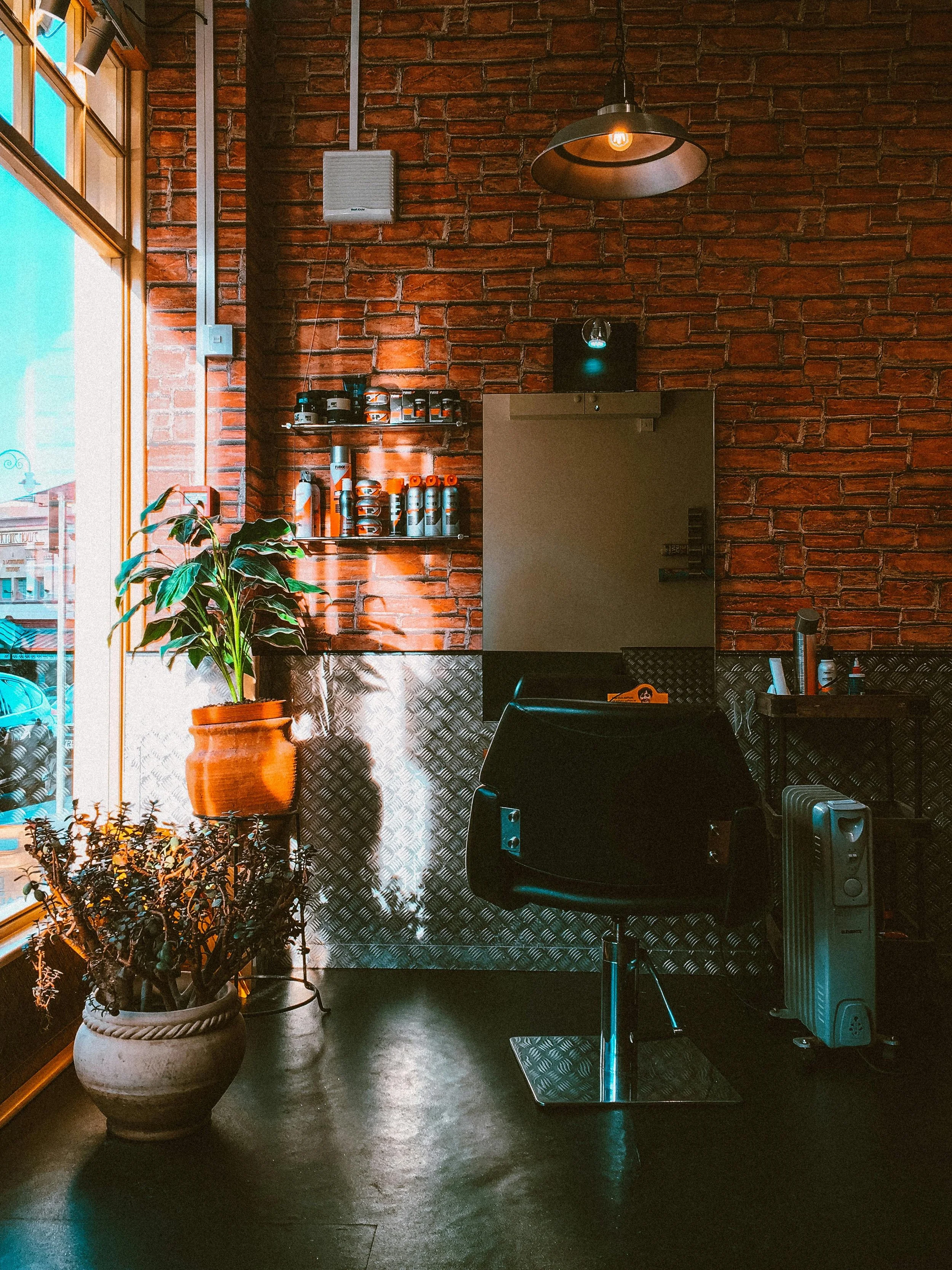 Interior of a modern barbershop with a barber chair, brick walls, plants, hair products on shelves, and a large window with natural light.
