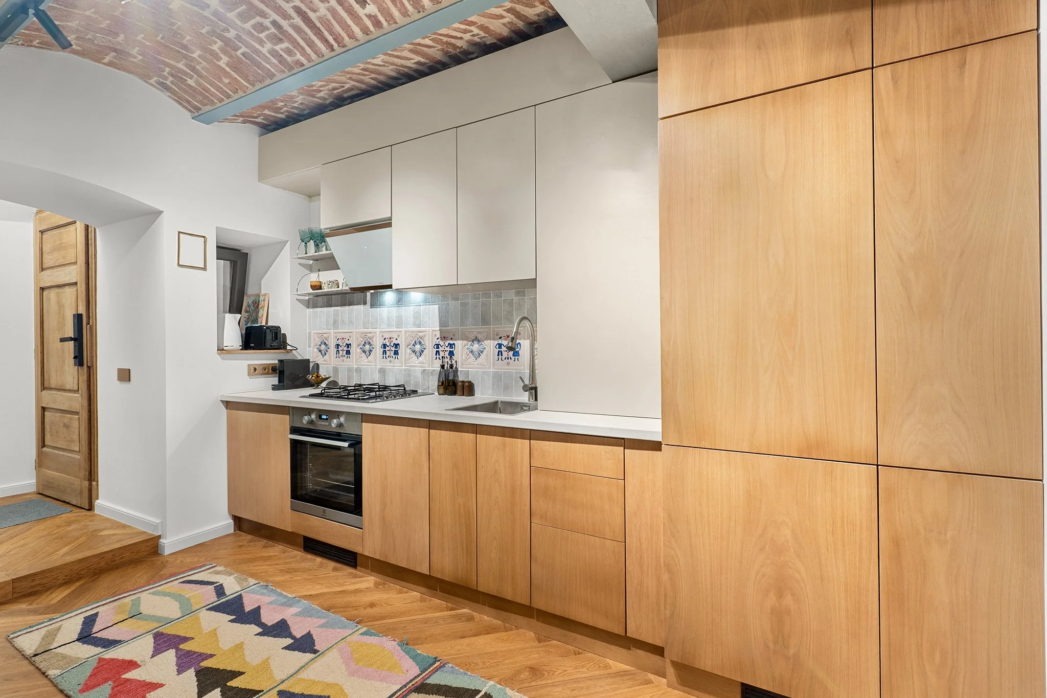Kitchen with wooden and white cabinets, a built-in oven, a gas stovetop, a small window, a colorful geometric rug, and a brick ceiling.