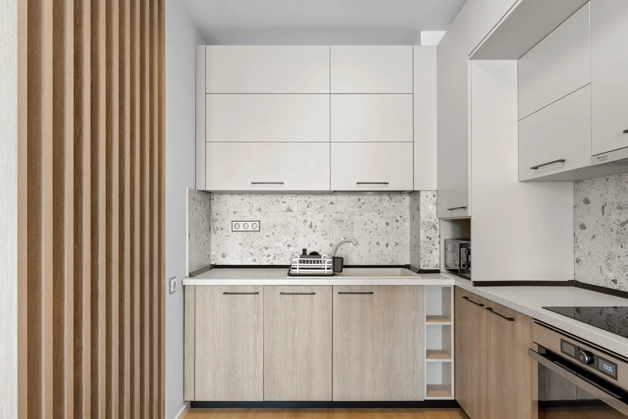 Modern kitchen with white cabinets, stone backsplash, wooden panel detail, built-in oven, and sink with dish rack.
