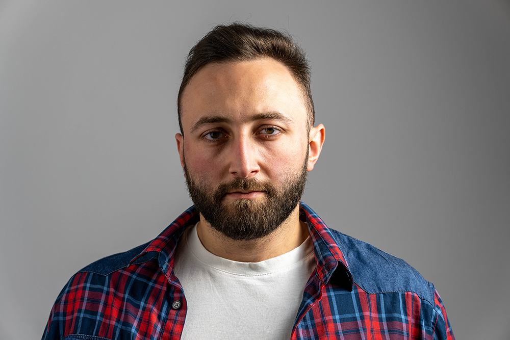 Man in plaid shirt and white t-shirt with neutral expression, against gray background.