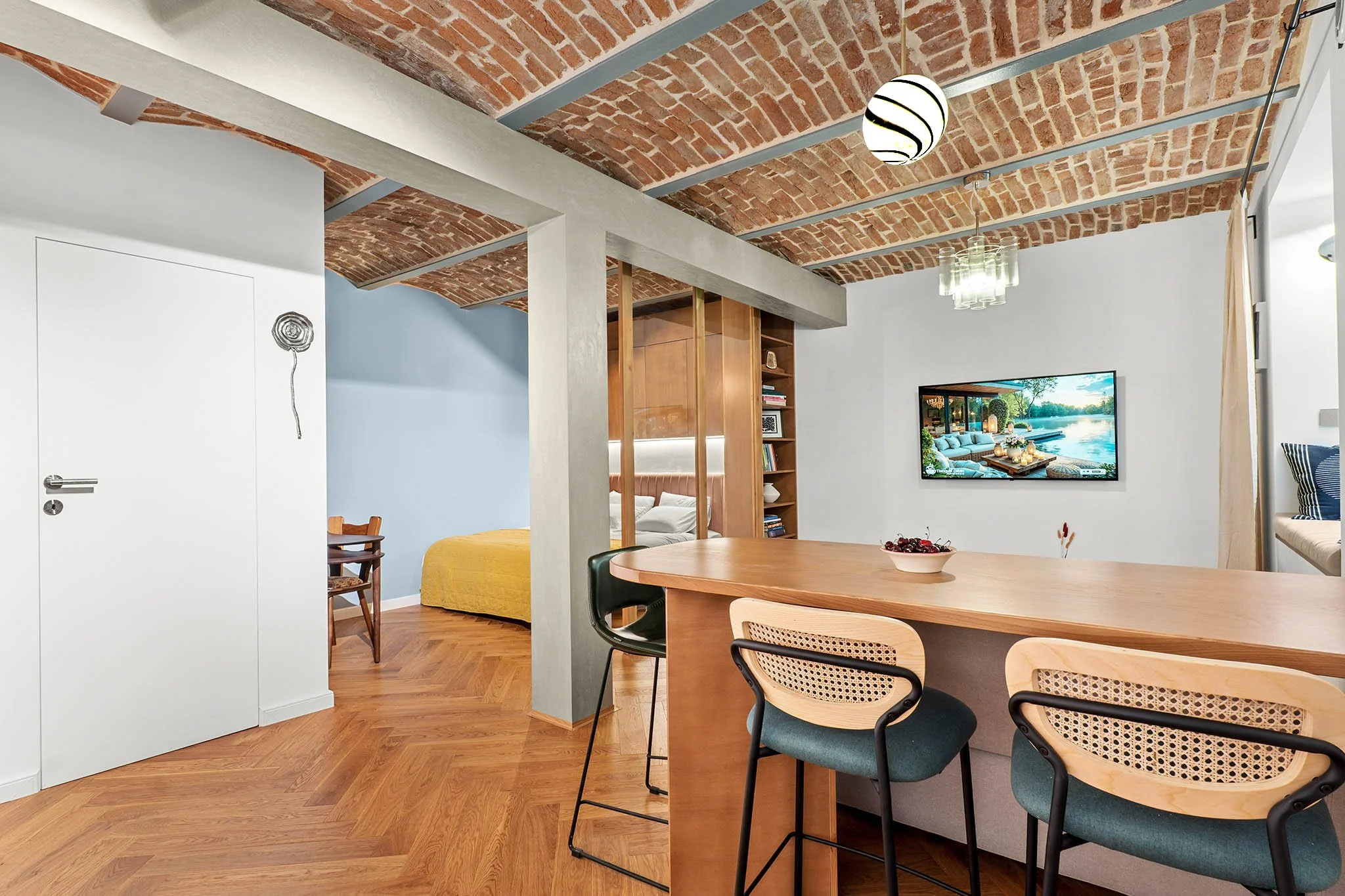 Open-concept living space with a dining area, a bedroom with a blue wall and a yellow bedspread, wooden flooring, exposed brick ceiling, TV on a white wall, and a window seat with striped pillows.