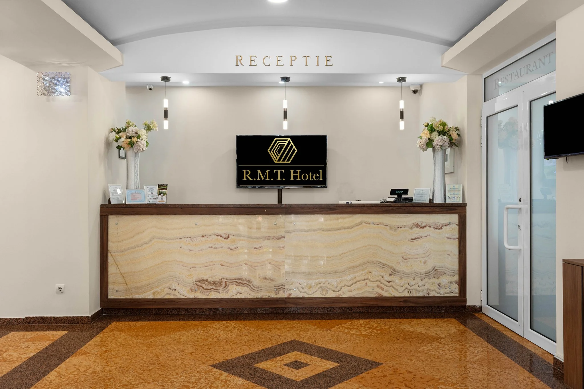 Hotel reception desk with a sign reading 'RECEPTION' and a television displaying the hotel name 'R.M.T. Hotel' above the counter. The counter is made of a marble-like material with a wooden frame, decorated with flower vases on either side. The backg
