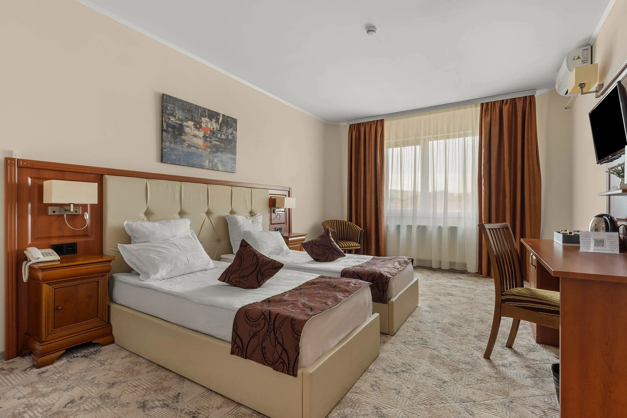 Hotel room with two single beds, brown and beige decor, a large window with sheer and brown curtains, a wooden desk and chair, a wall-mounted TV, and paintings on the wall.