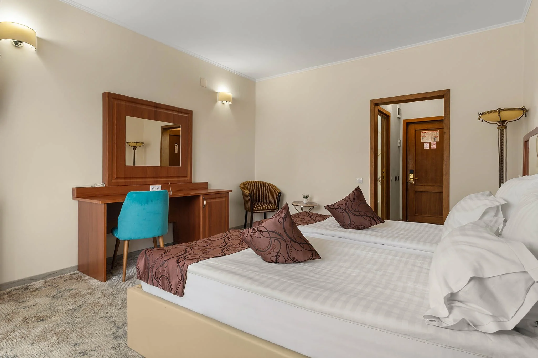 A hotel room with two twin beds, white linens, brown decorative pillows, a wooden dresser with a mirror, a blue chair, and a small side table with a plant. The room has beige walls, two wall-mounted lamps, and a wooden door.