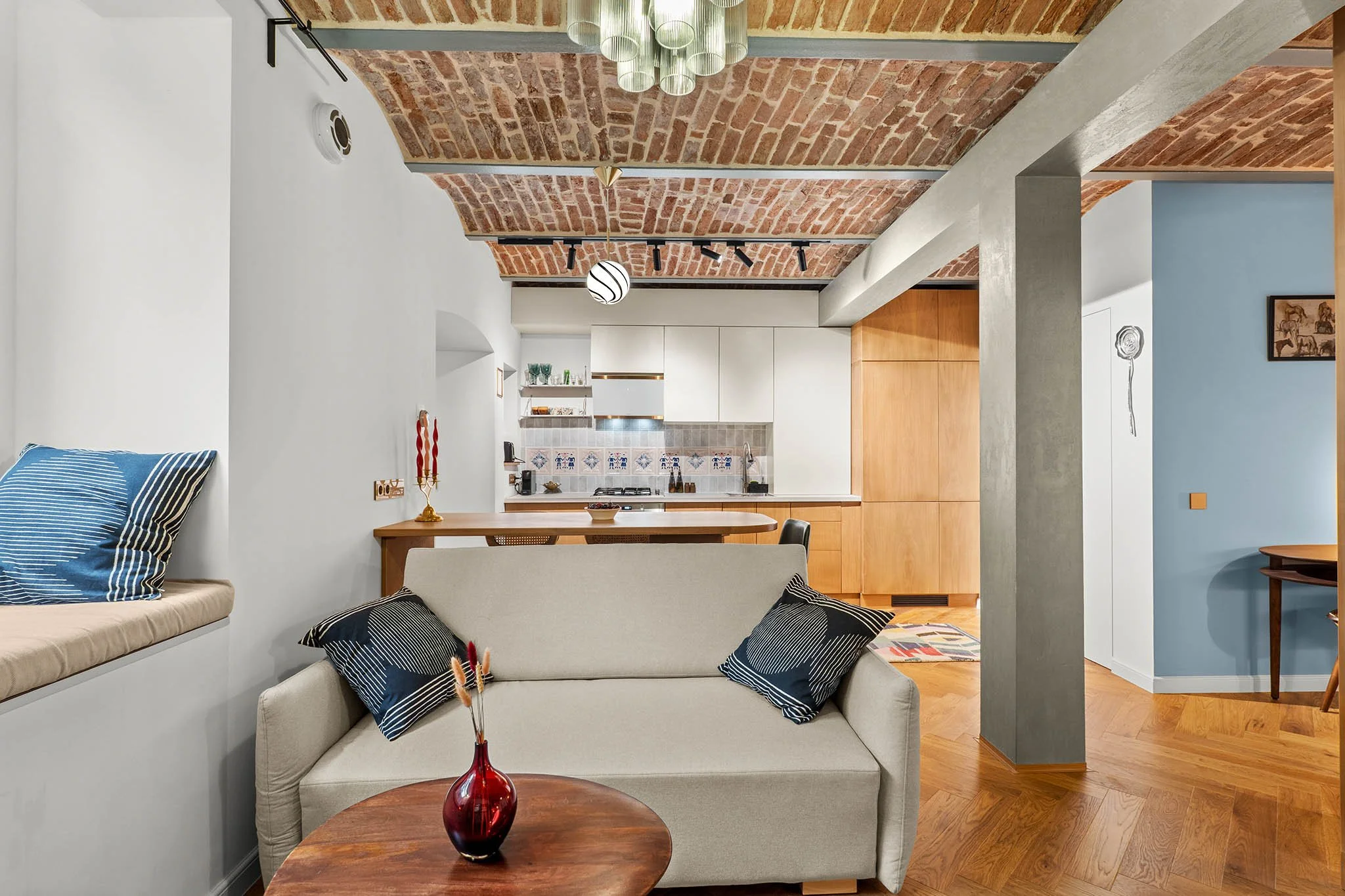 Living room with a beige sofa and black and white patterned pillows, a round wooden coffee table with a red vase, and a built-in window seat with blue striped cushion, overlooking an open kitchen with white cabinets and wooden accents, exposed brick 