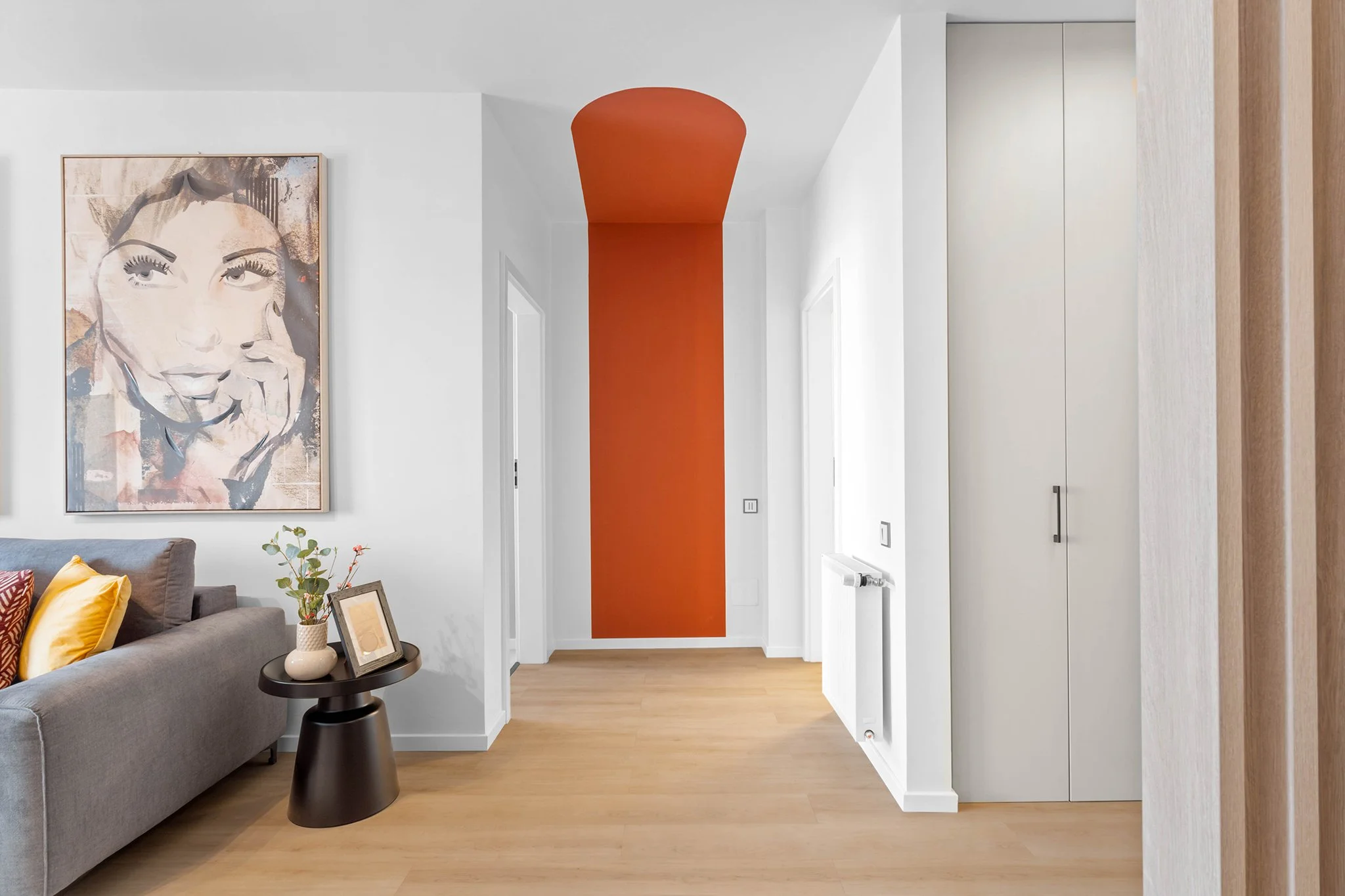 Modern living room with a gray sofa, colorful cushions, and a side table with a vase and frame. A large portrait is on the wall. An adjacent hallway features an orange accent wall and ceiling with light wooden flooring.